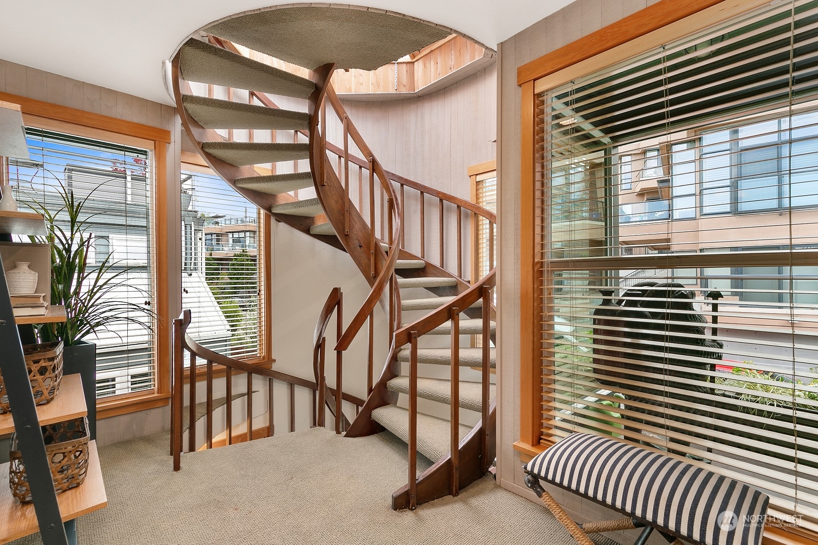 Curved wooden staircase with carpeted steps, large blinded windows, and a striped bench on the landing.