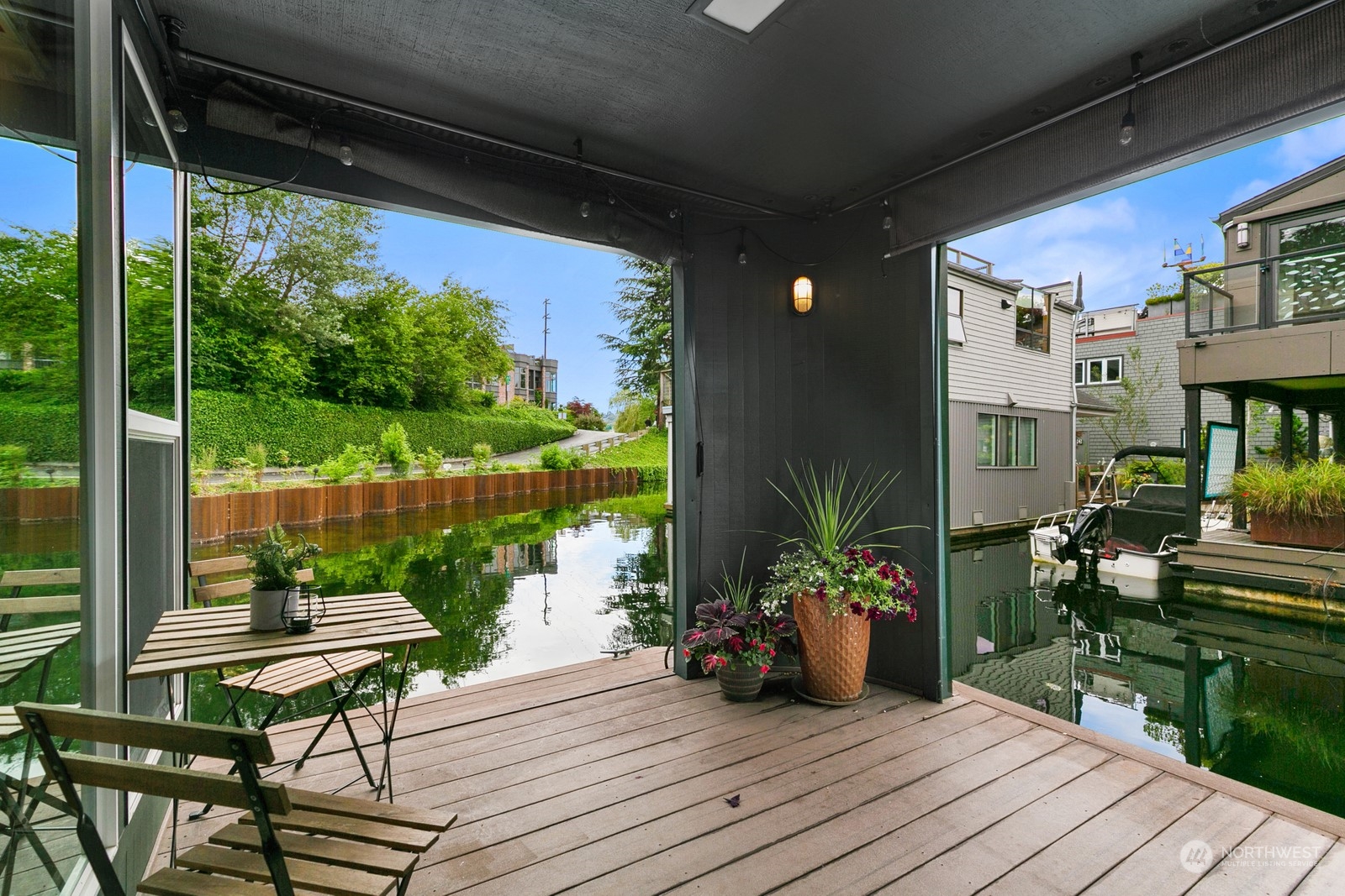 Covered wood deck beside a canal, with outdoor seating, potted flowers, and neighboring homes across the water.