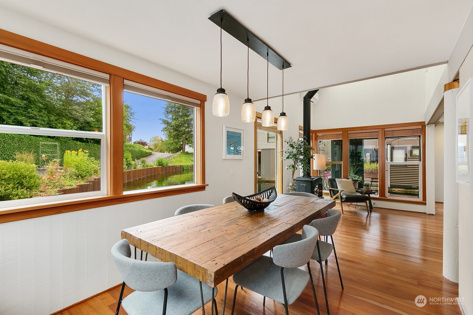 Dining room with a rustic wood table, pendant lights, hardwood floors, and large windows overlooking greenery.