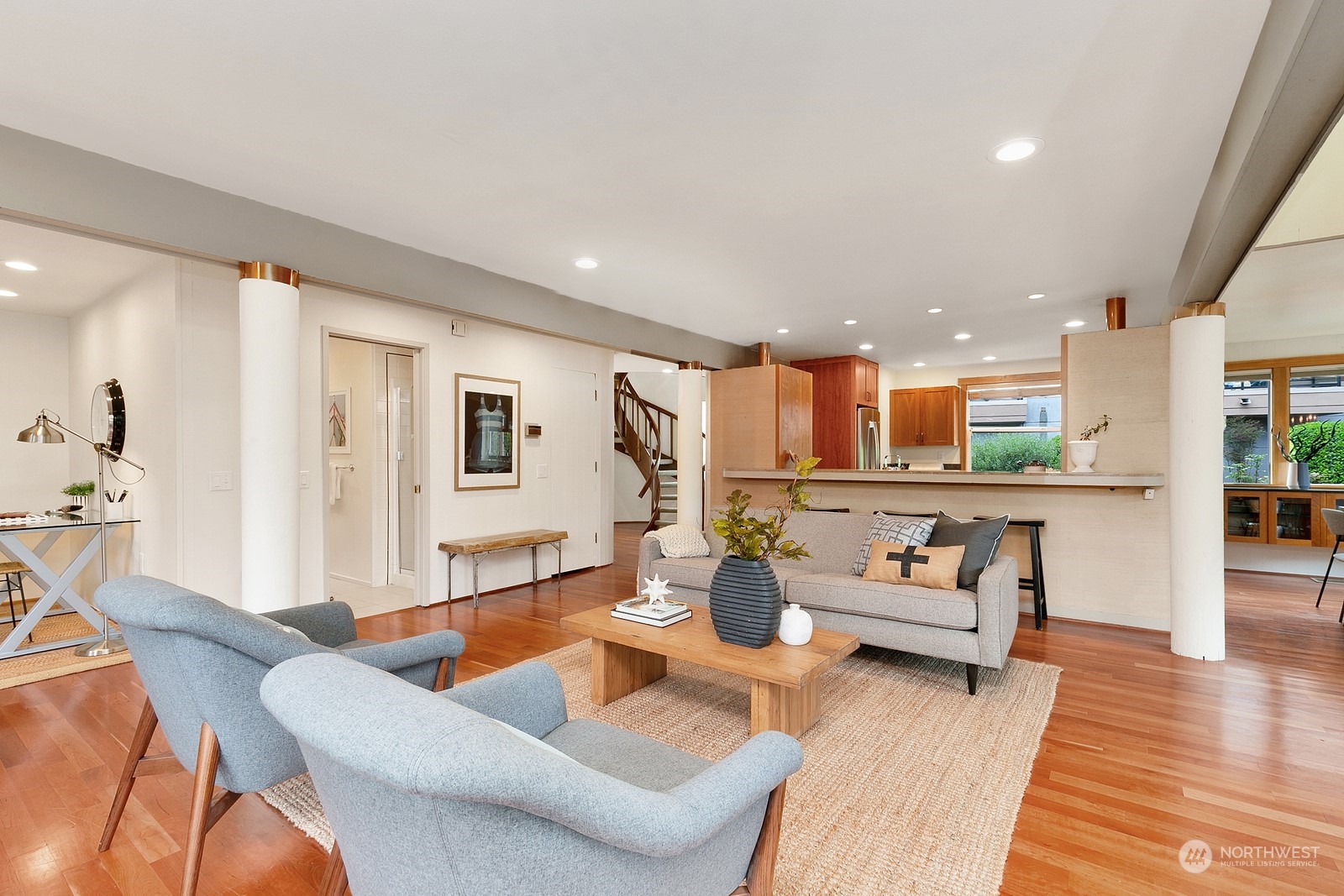 Staged living room with blue armchairs, gray sofa, hardwood floors, and an adjoining kitchen.