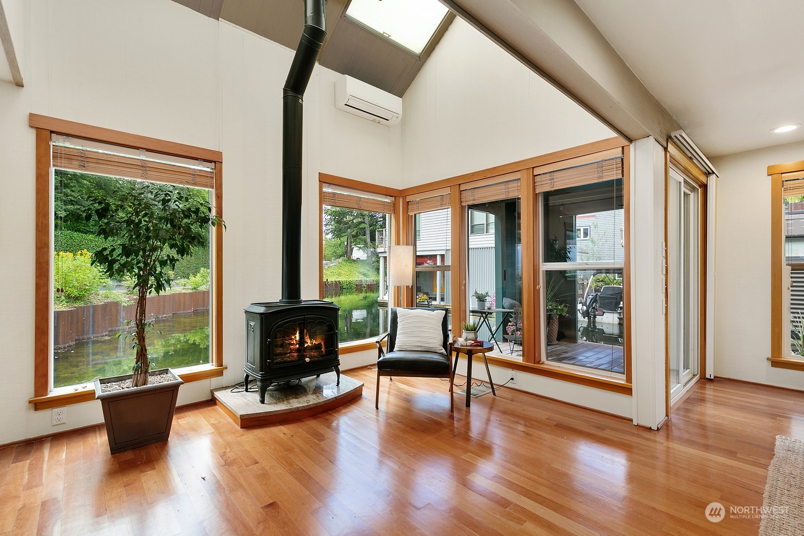 Living room with a black wood stove, vaulted ceiling, and large windows overlooking water.