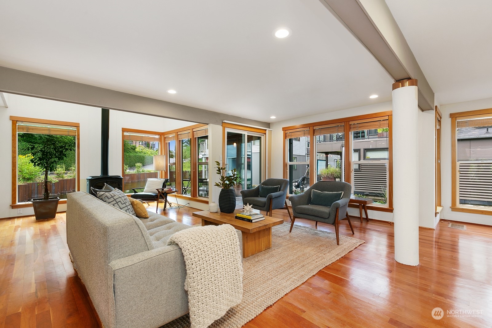 Living room with a wood-burning stove, gray seating, hardwood floors, and wide windows.