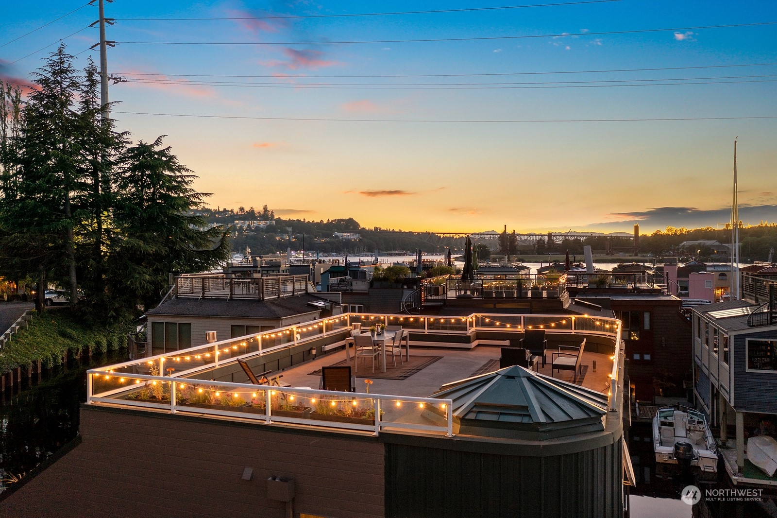 Rooftop deck with string lights and seating overlooking a marina at sunset.