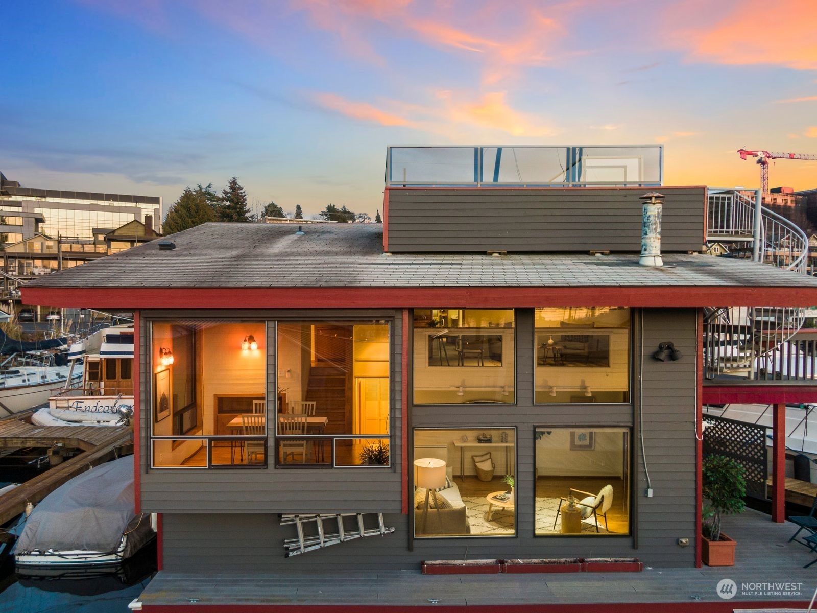 Modern waterfront home with glowing windows, rooftop terrace, and boats moored alongside.