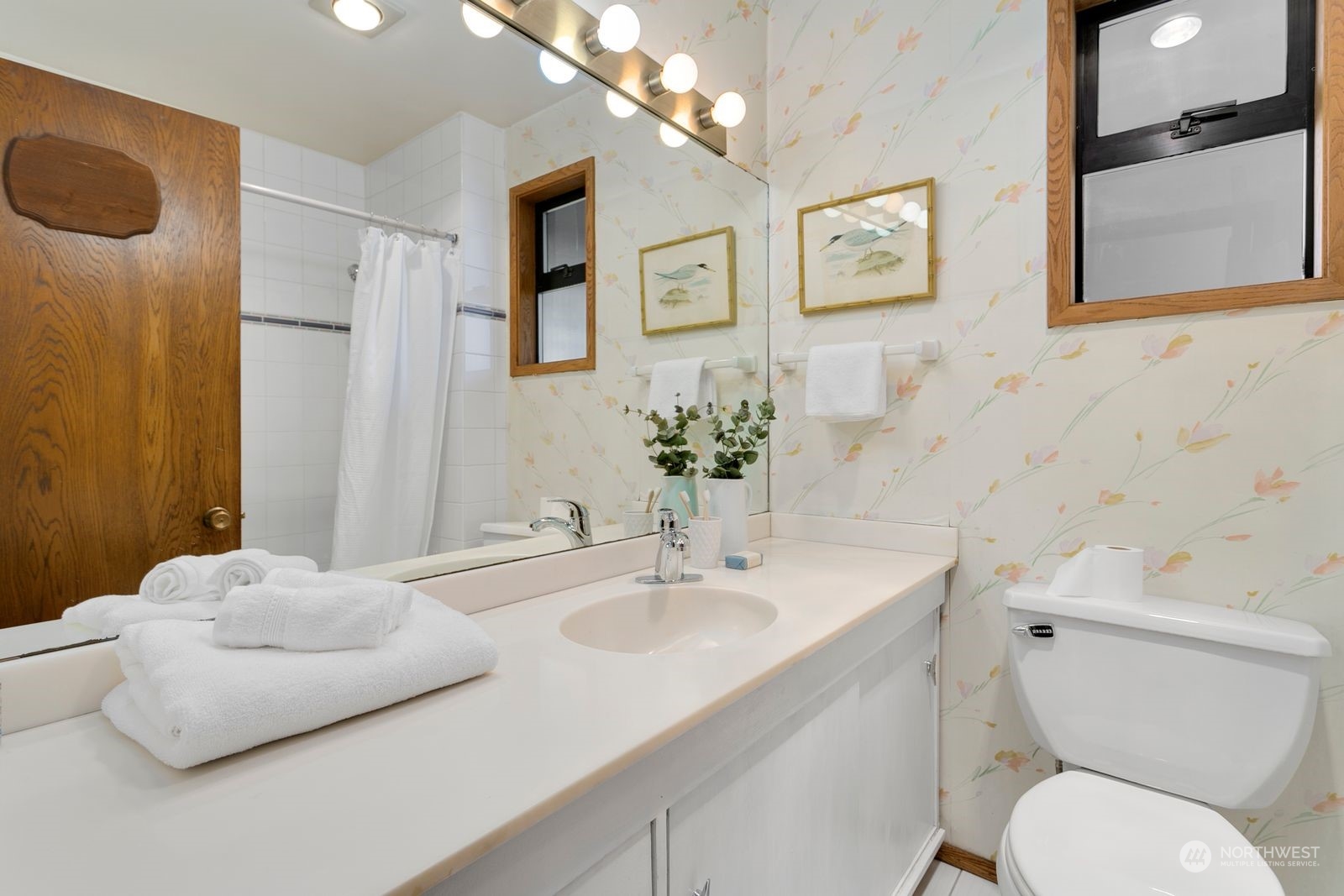 Bathroom with a long white vanity, floral wallpaper, and a tiled shower.