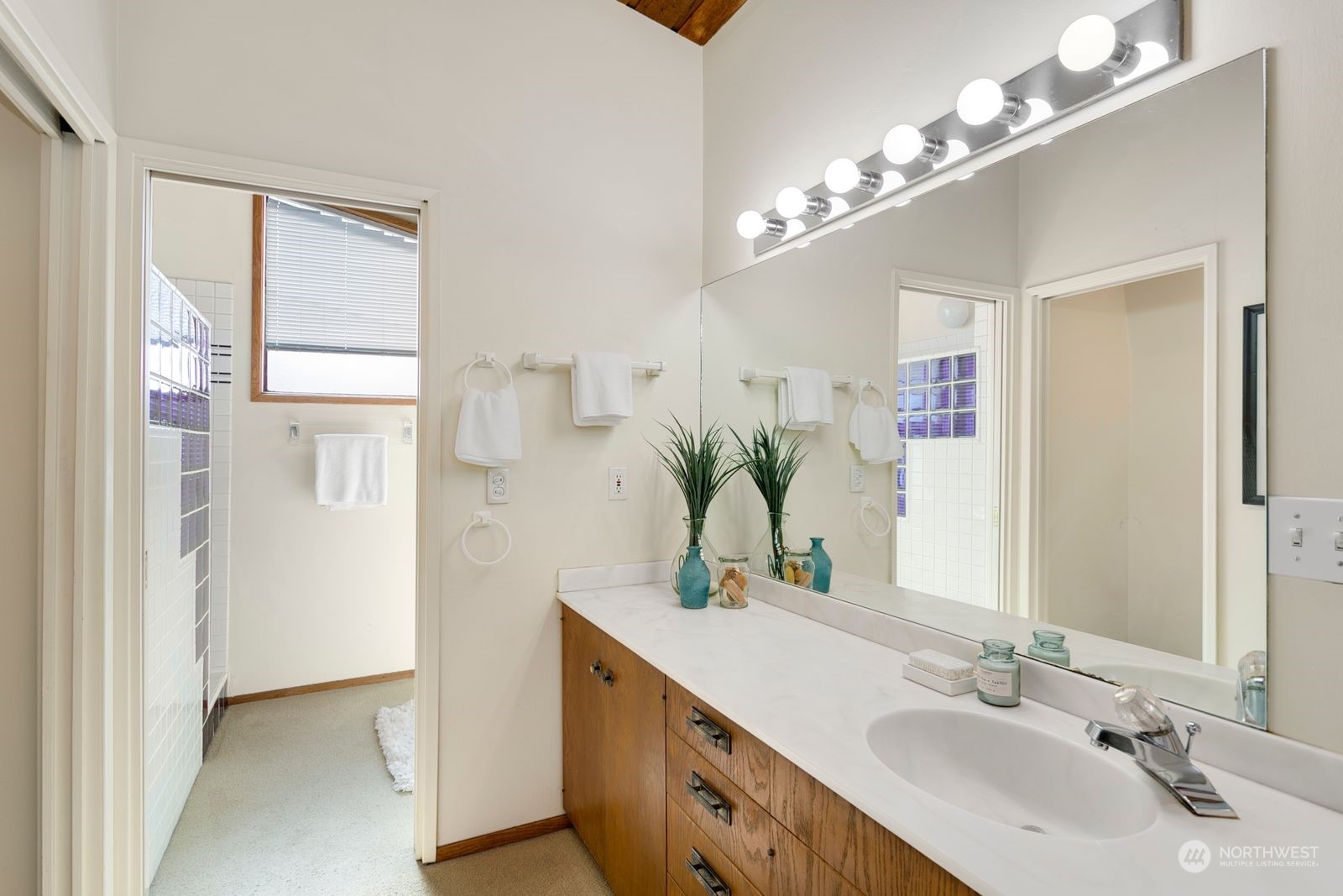 Bathroom with a long wood-cabinet vanity, large lit mirror, and glass-block shower accents.