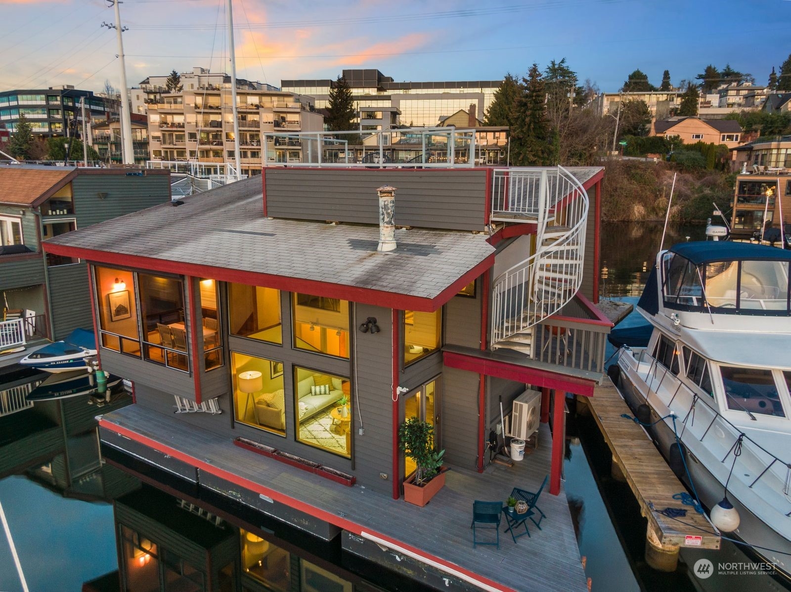 Modern gray floating home with red trim, rooftop spiral staircase, and boats moored alongside at sunset.
