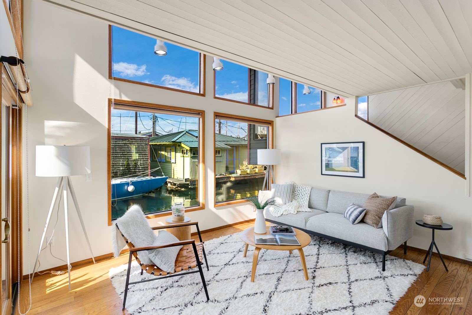 Living room with vaulted ceiling, large windows overlooking boats and waterfront homes, and a gray sofa.