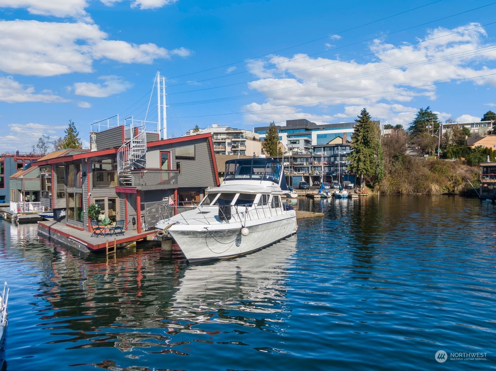 Floating home on the water beside a moored white yacht, with urban buildings in the background.