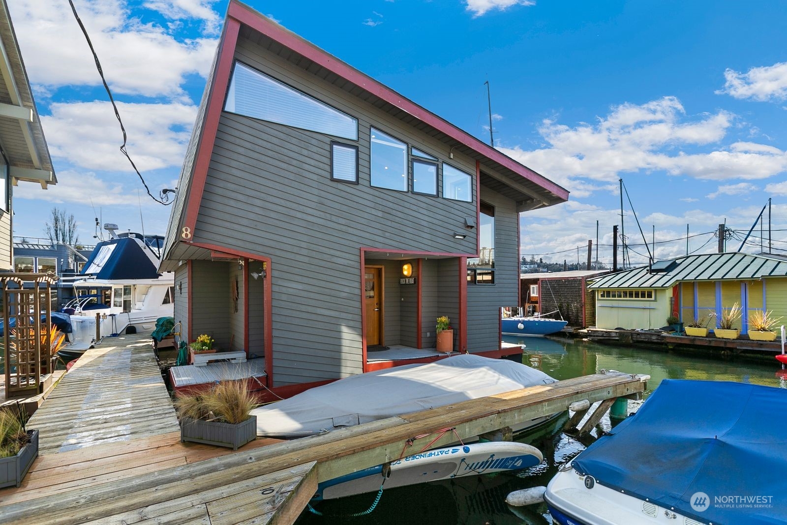 Gray floating home with red trim and a steep roof, moored beside boats and wooden docks.