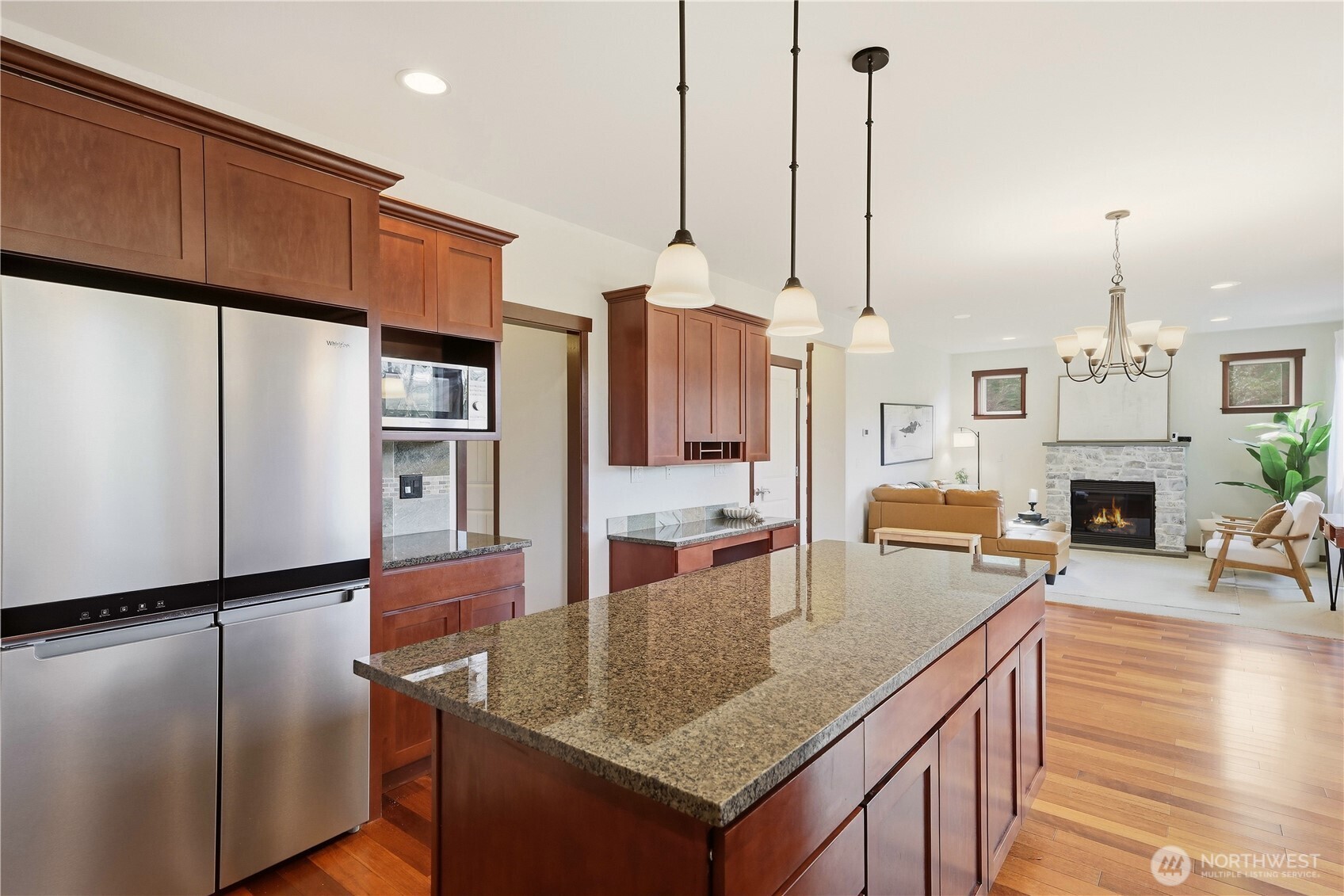 Kitchen with a granite island and stainless steel appliances opening to a living room with a stone fireplace.
