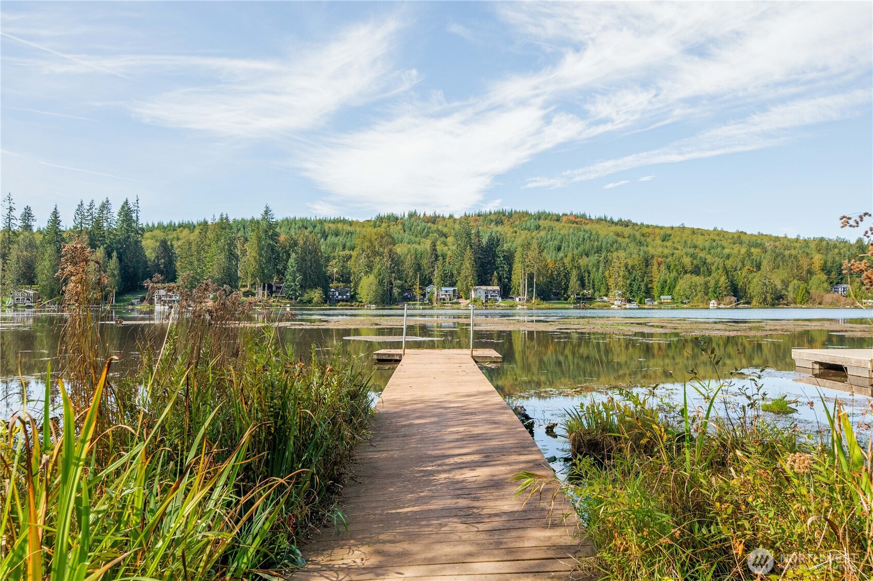 Wooden dock reaches into a calm lake, with shoreline homes and a forested hillside across the water.