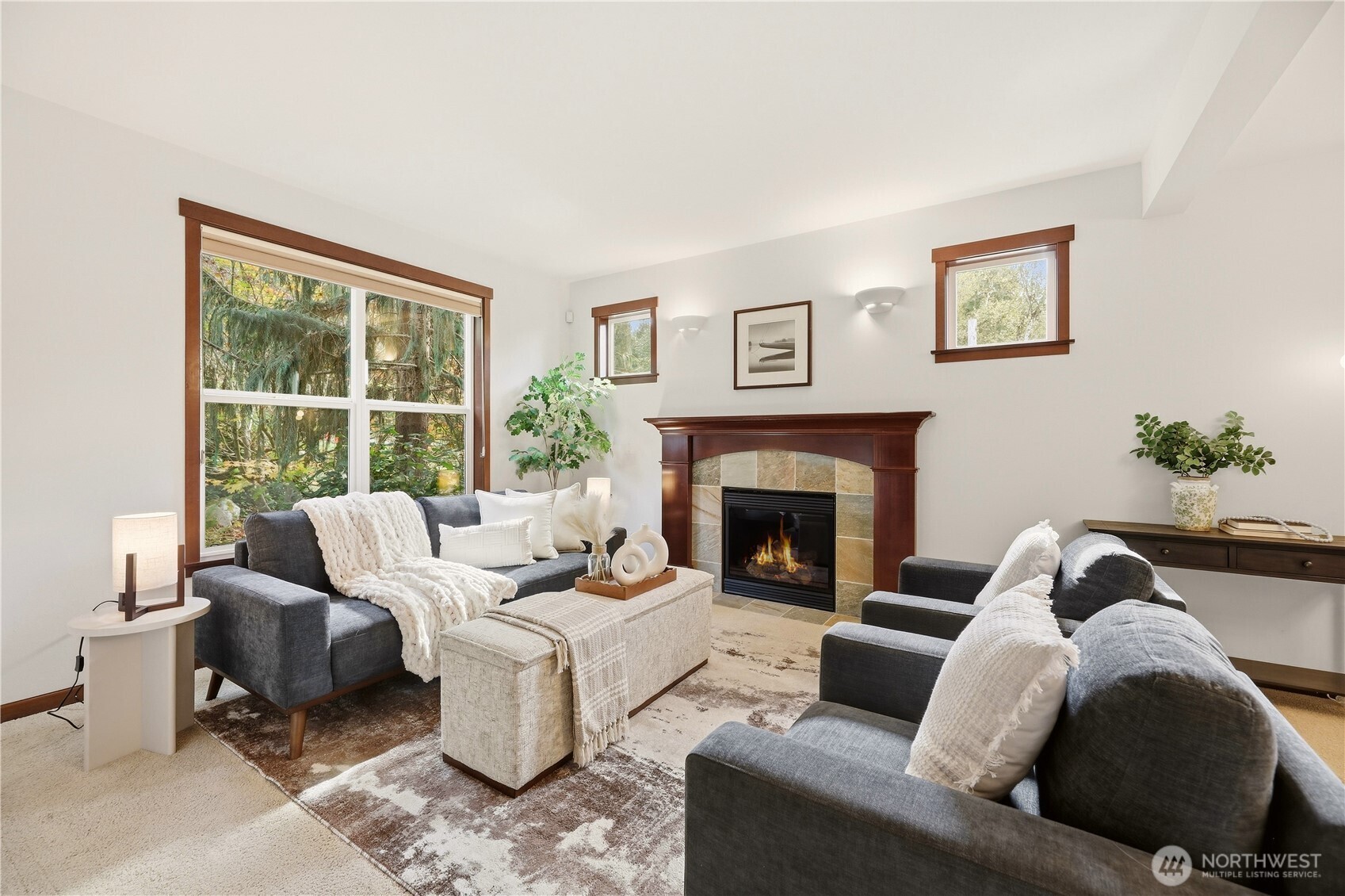 Staged living room with charcoal sofas, a stone fireplace, and large windows overlooking trees.