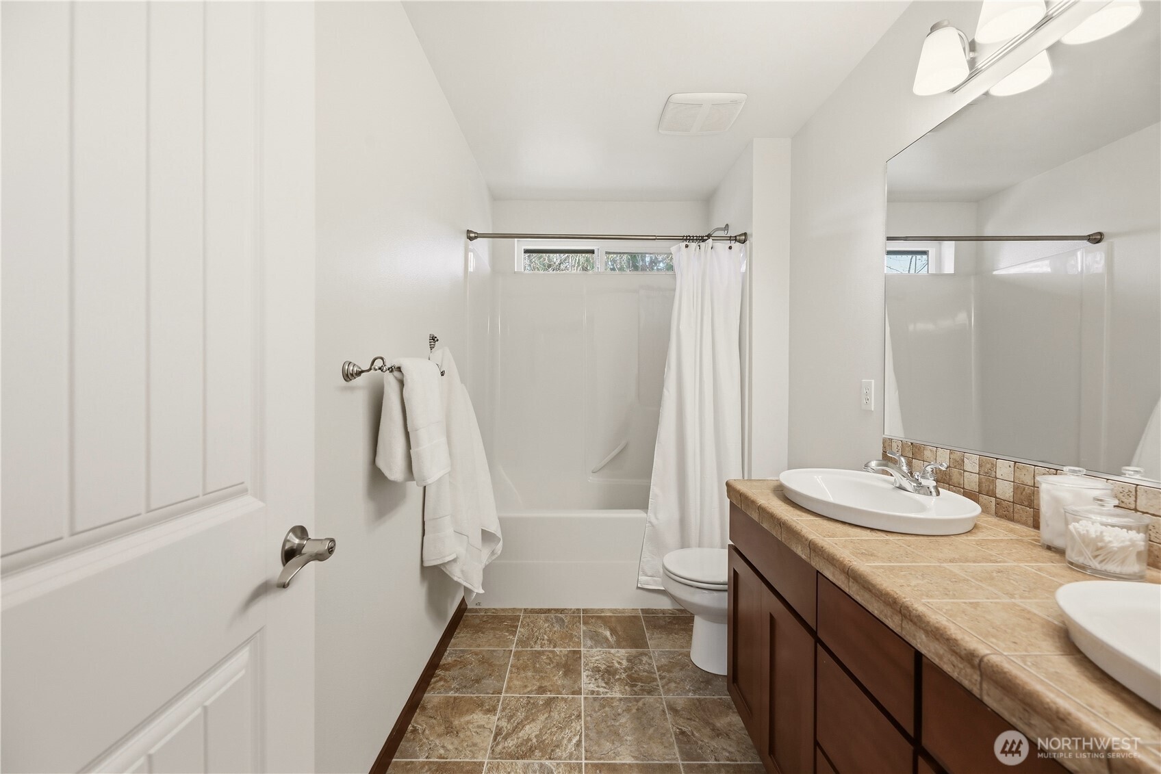 Bathroom with a white tub and shower, brown tiled floor, and long double-sink vanity.
