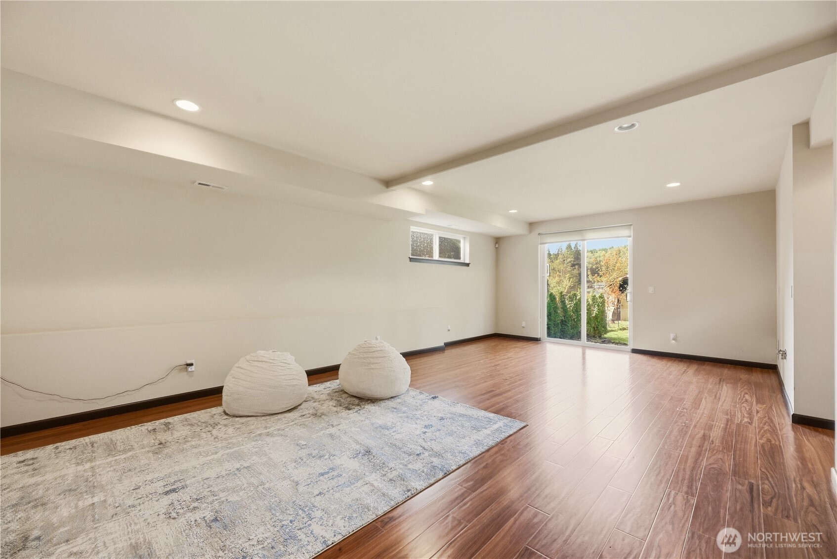 Living area with wood-look floors, a large rug, two poufs, and sliding glass doors to the garden.