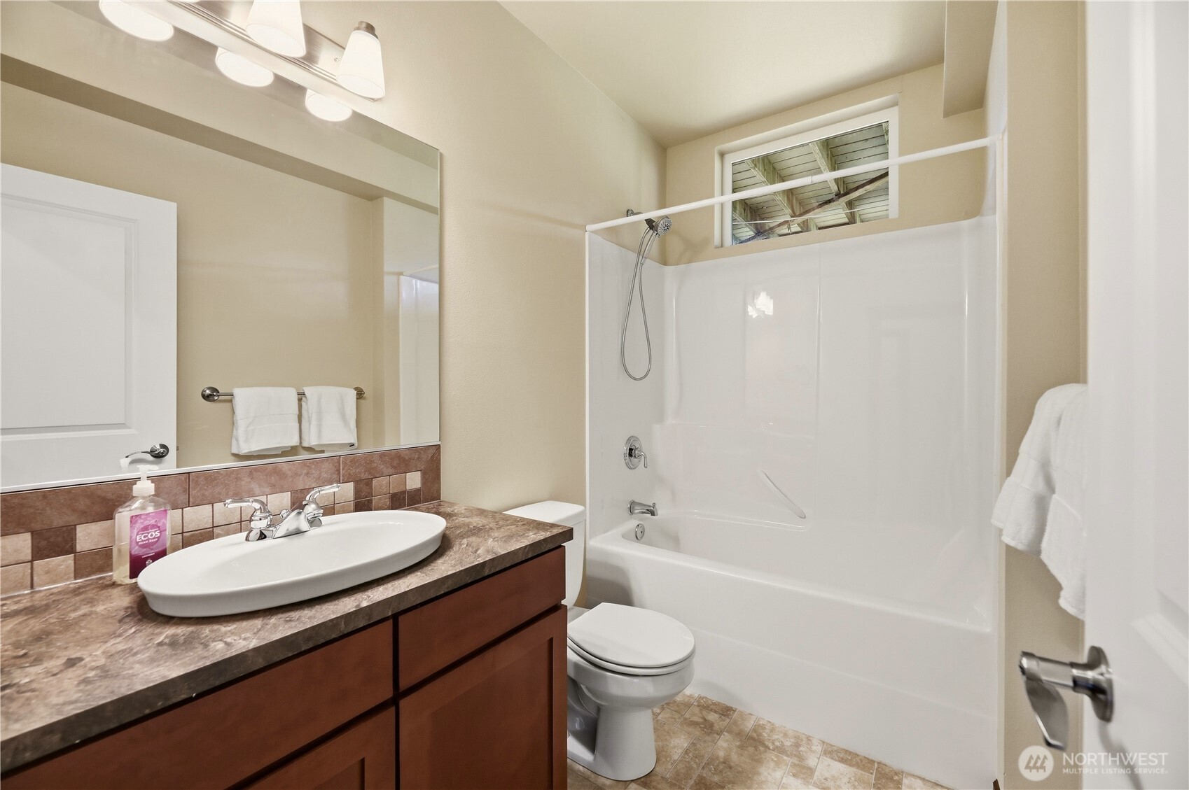 Bathroom with a brown tiled vanity, white tub and shower, and a high window above the tub.
