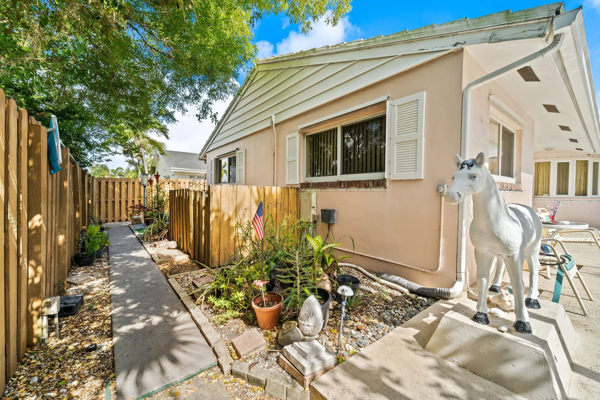 House exterior beside a fenced garden walkway, with potted plants and a white horse statue.