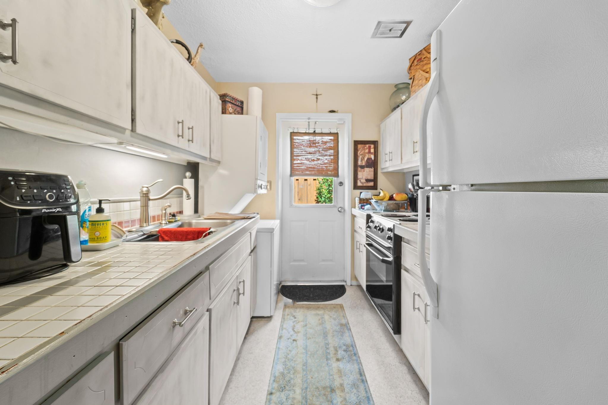 Galley kitchen with white cabinets, tiled counters, and a rear door with a bamboo blind.