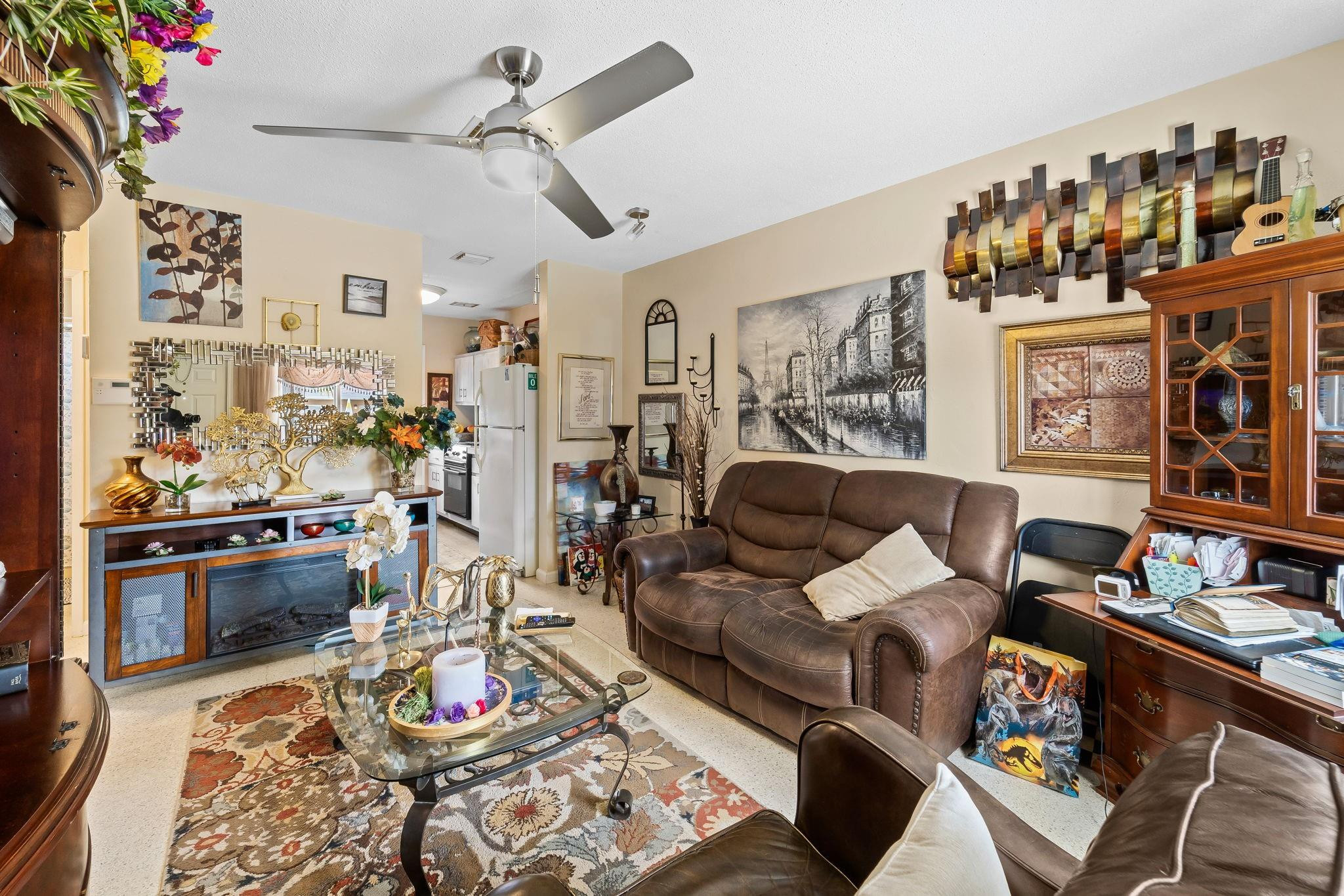 Furnished living room with brown leather sofas, glass coffee table, patterned rug, and kitchen beyond.