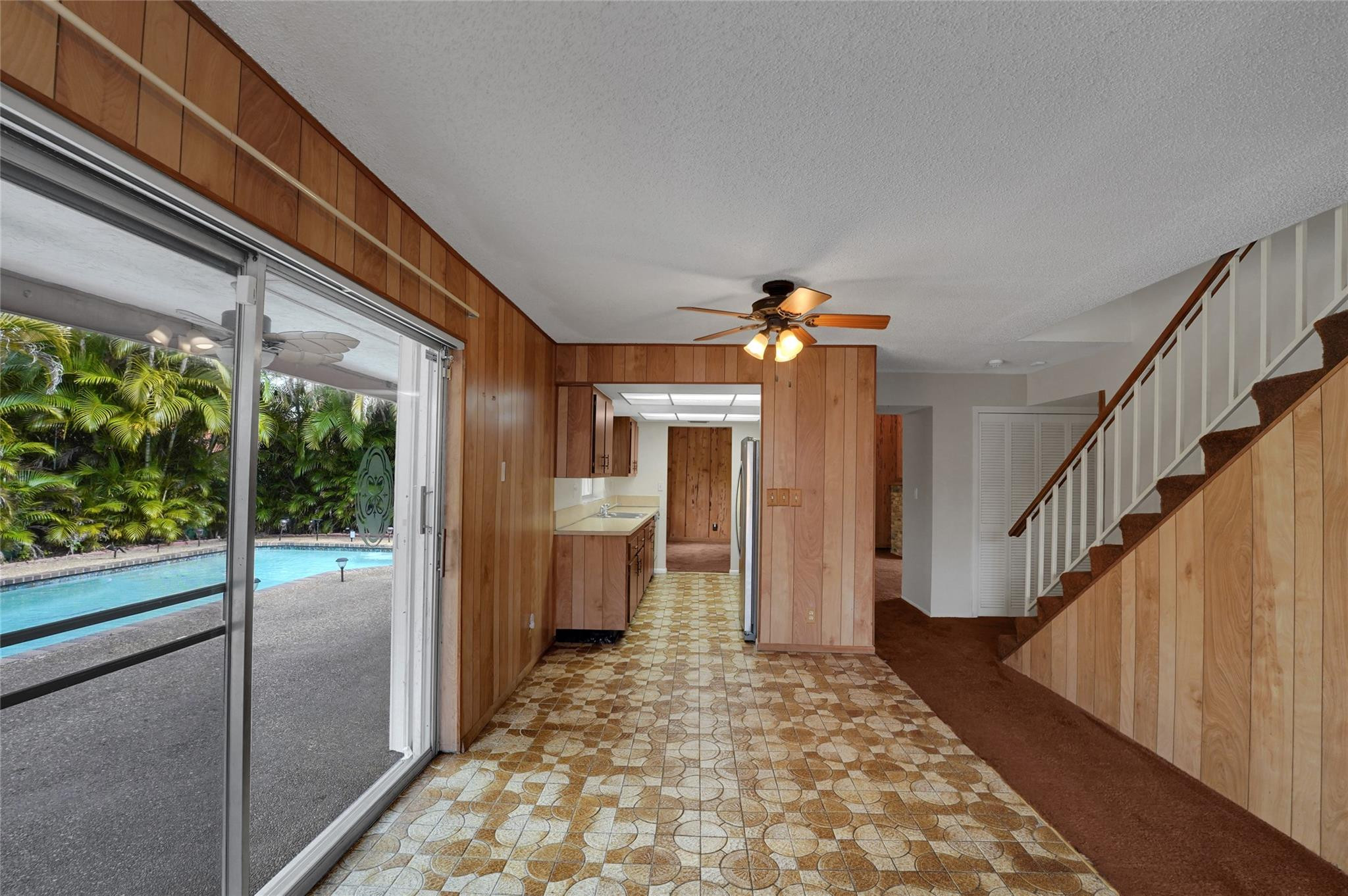 Wood-paneled living area with patterned tile, staircase, galley kitchen, and sliding doors overlooking a pool.