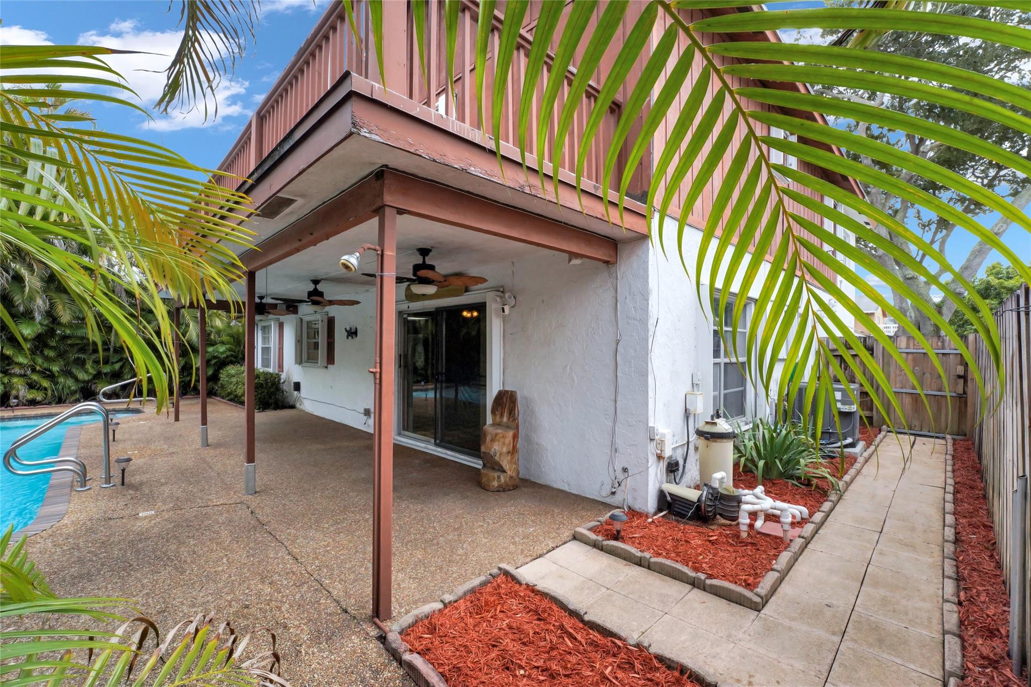 Covered backyard patio beside a swimming pool, with a second-story balcony and tropical palms.