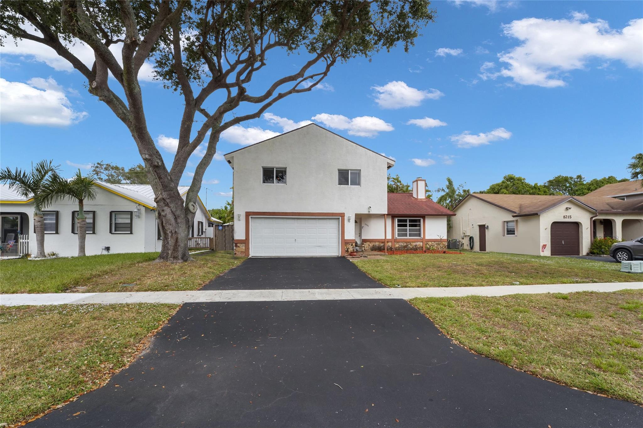 Two-story white stucco house with a double garage, broad driveway, and large front-yard tree.