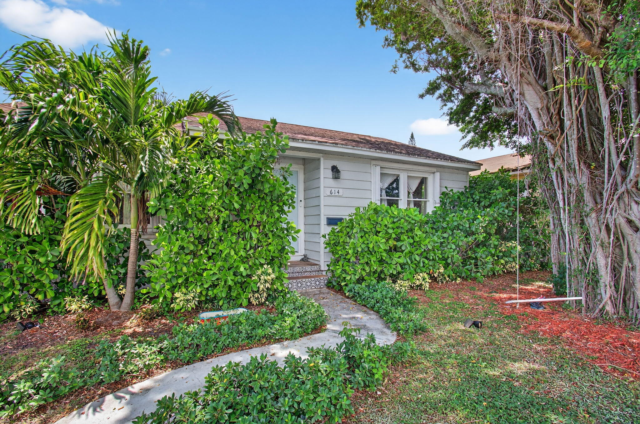 Single-story gray house with a tropical, shrub-lined walkway and a large banyan tree.