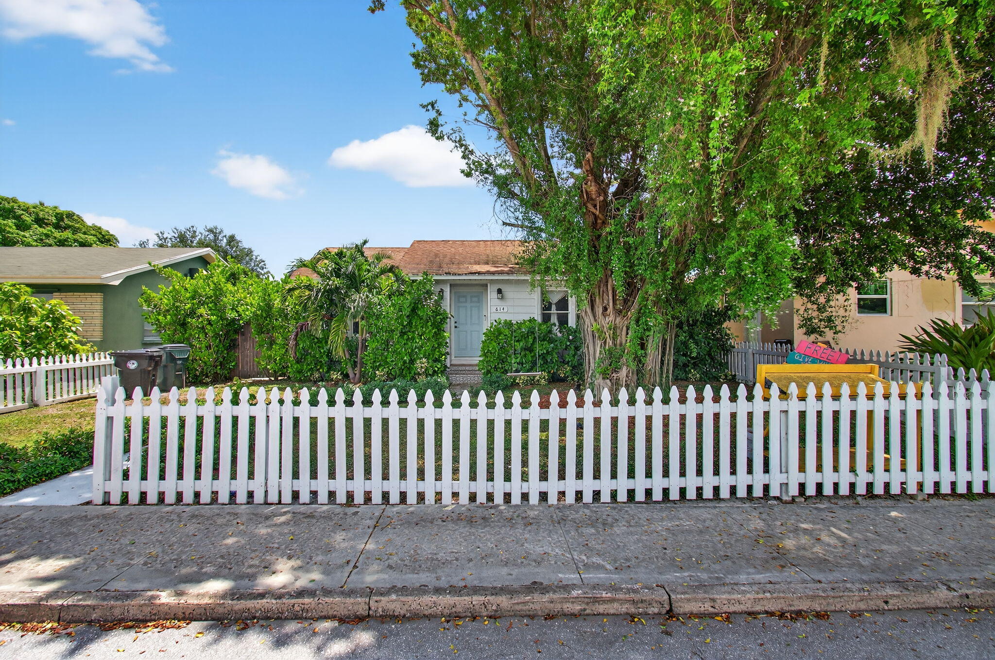 Small white house with a blue door behind a white picket fence and large shade tree.