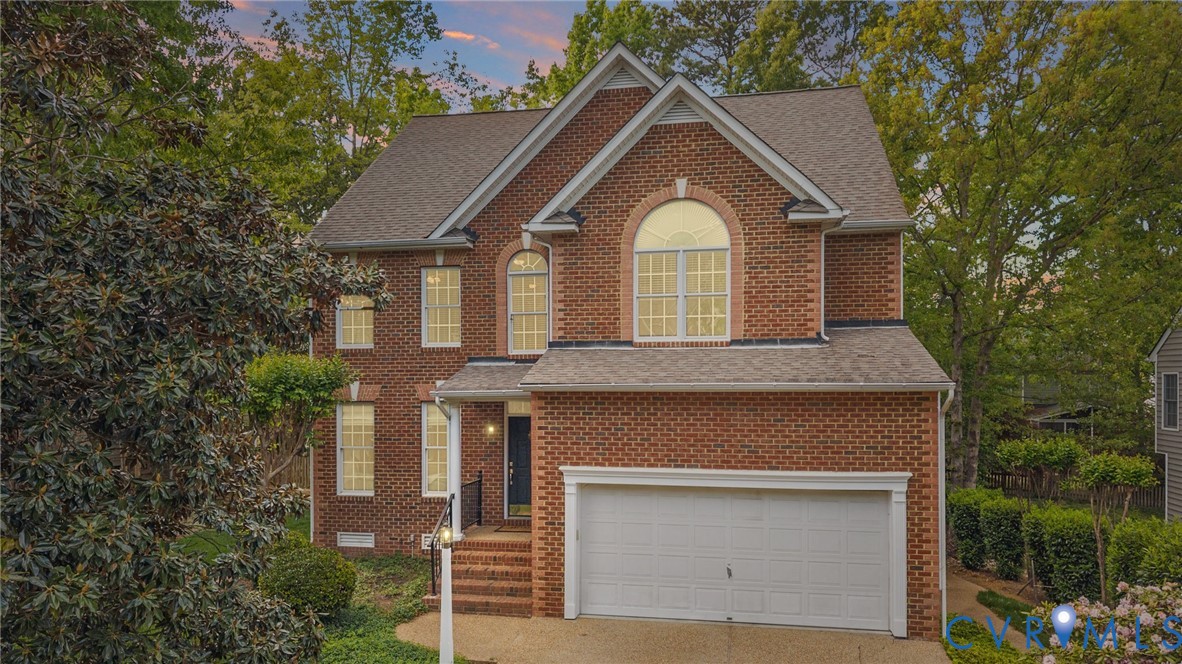 Two-story brick house with a white double garage, arched upper window, and mature trees.