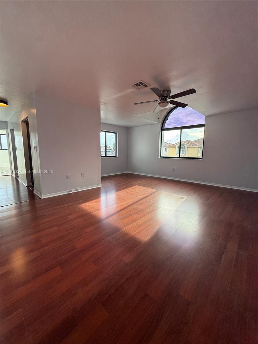 Empty living room with reddish-brown hardwood floors, ceiling fan, and large arched window.
