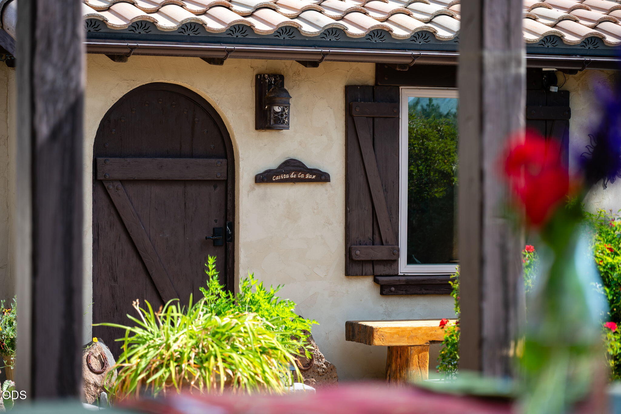 Stucco home entrance with an arched wooden door, tiled roof, and garden plants.