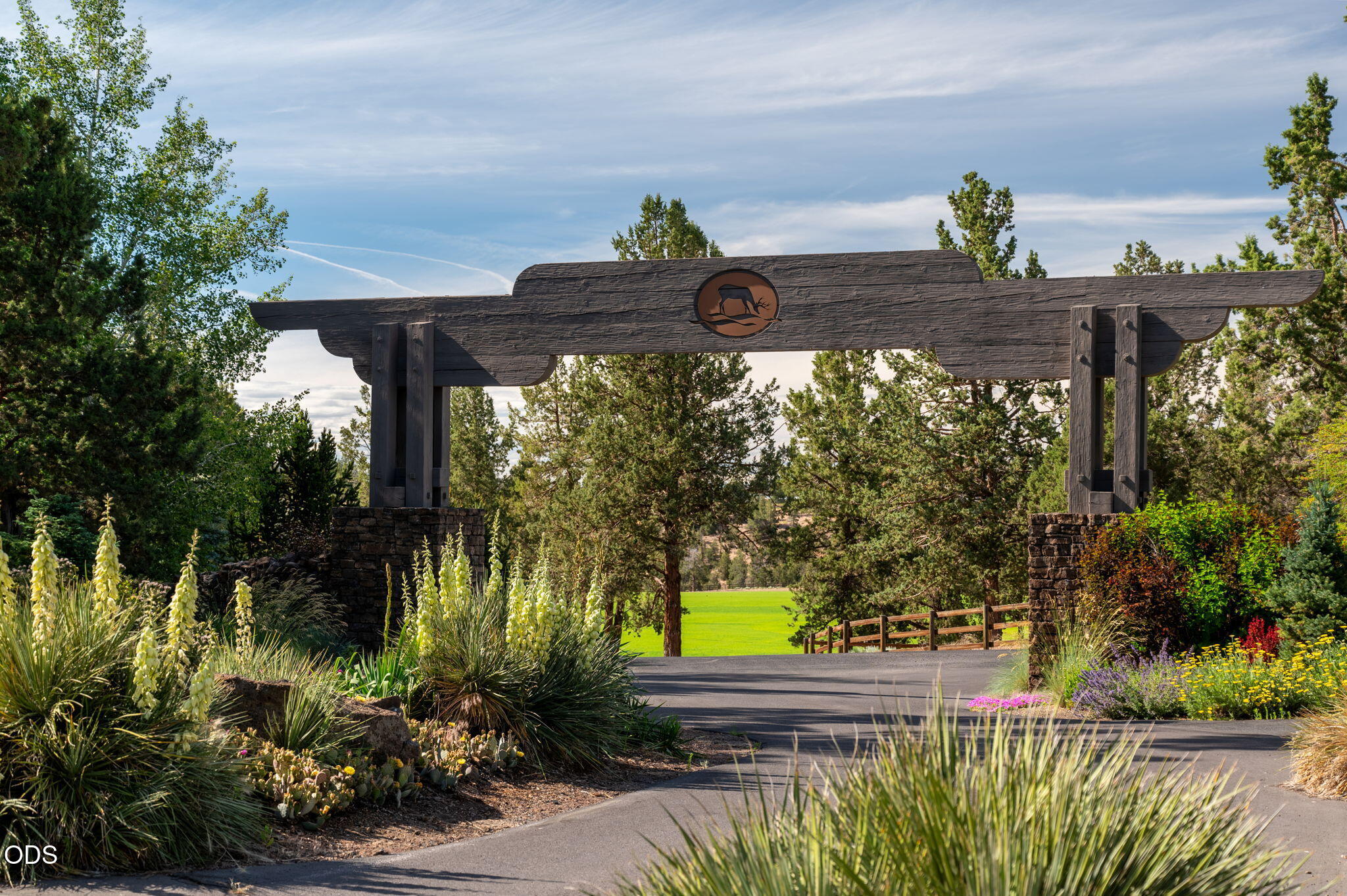 Landscaped entrance gateway with dark timber beams, stone pillars, and a paved road.