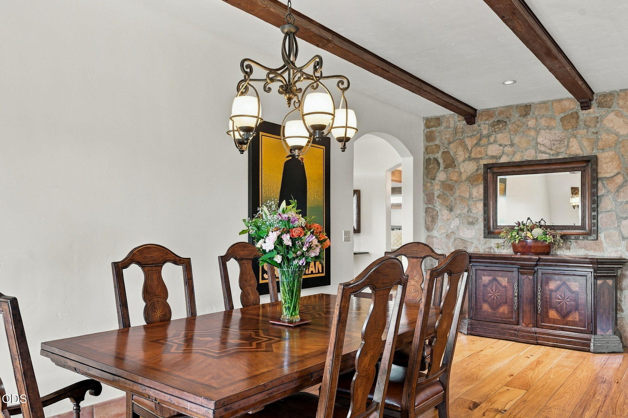 Dining room with a carved wood table, brass chandelier, exposed beams, and stone accent wall.