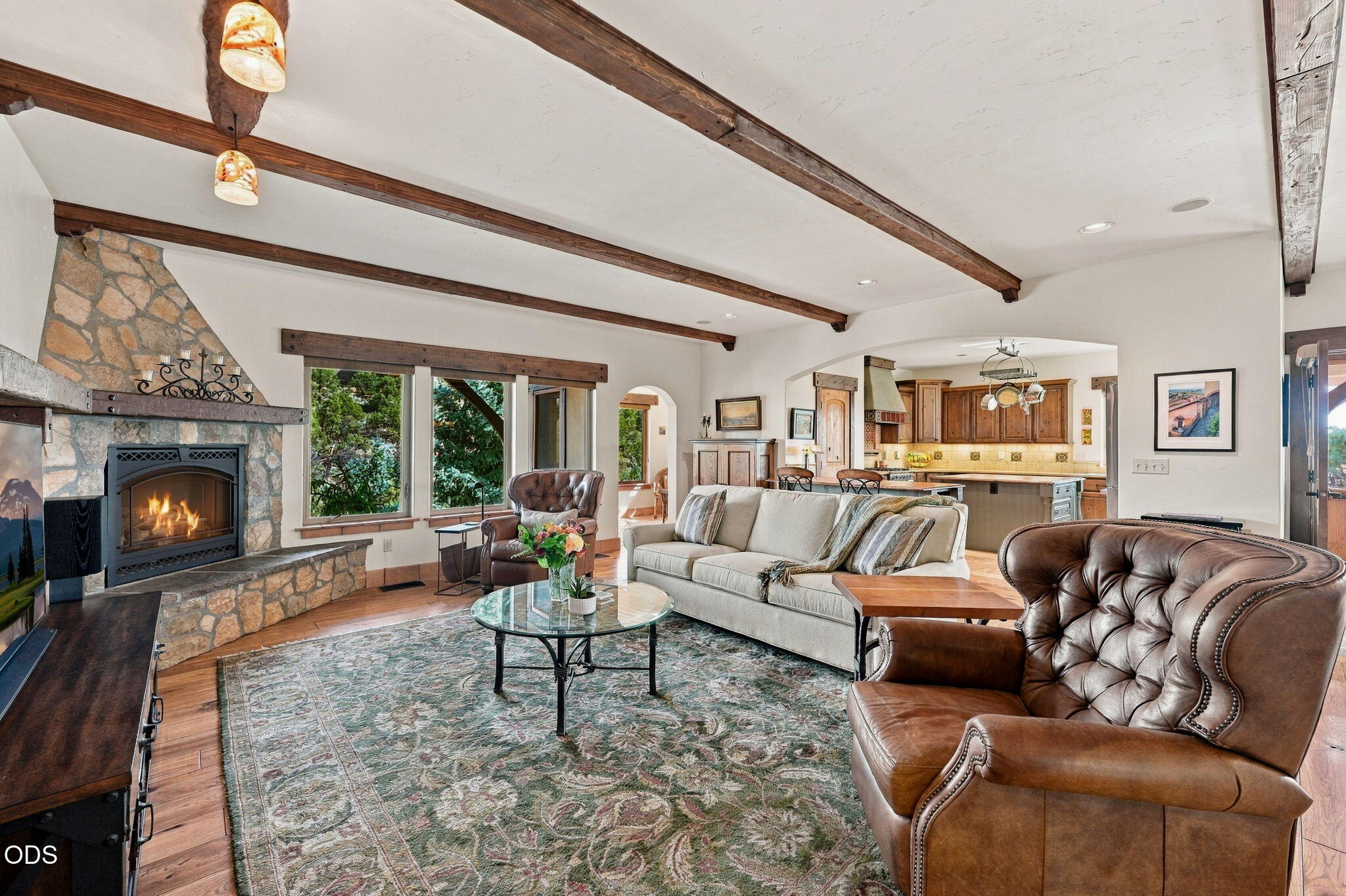 Living room with a stone fireplace, exposed wood beams, leather chairs, and an open kitchen