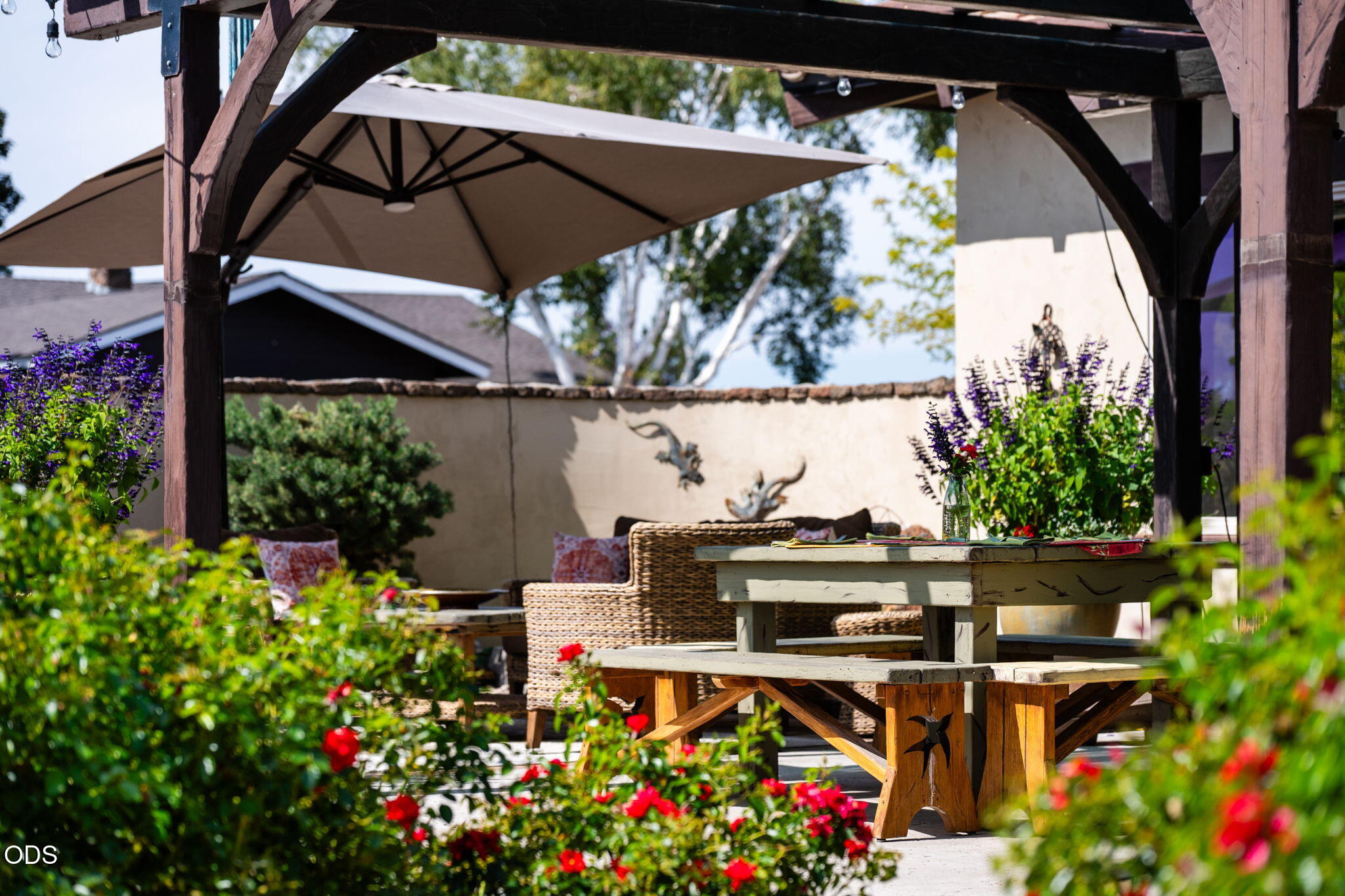 Outdoor patio with a timber pergola, shade umbrella, rustic dining table, wicker seating, and flowering garden.