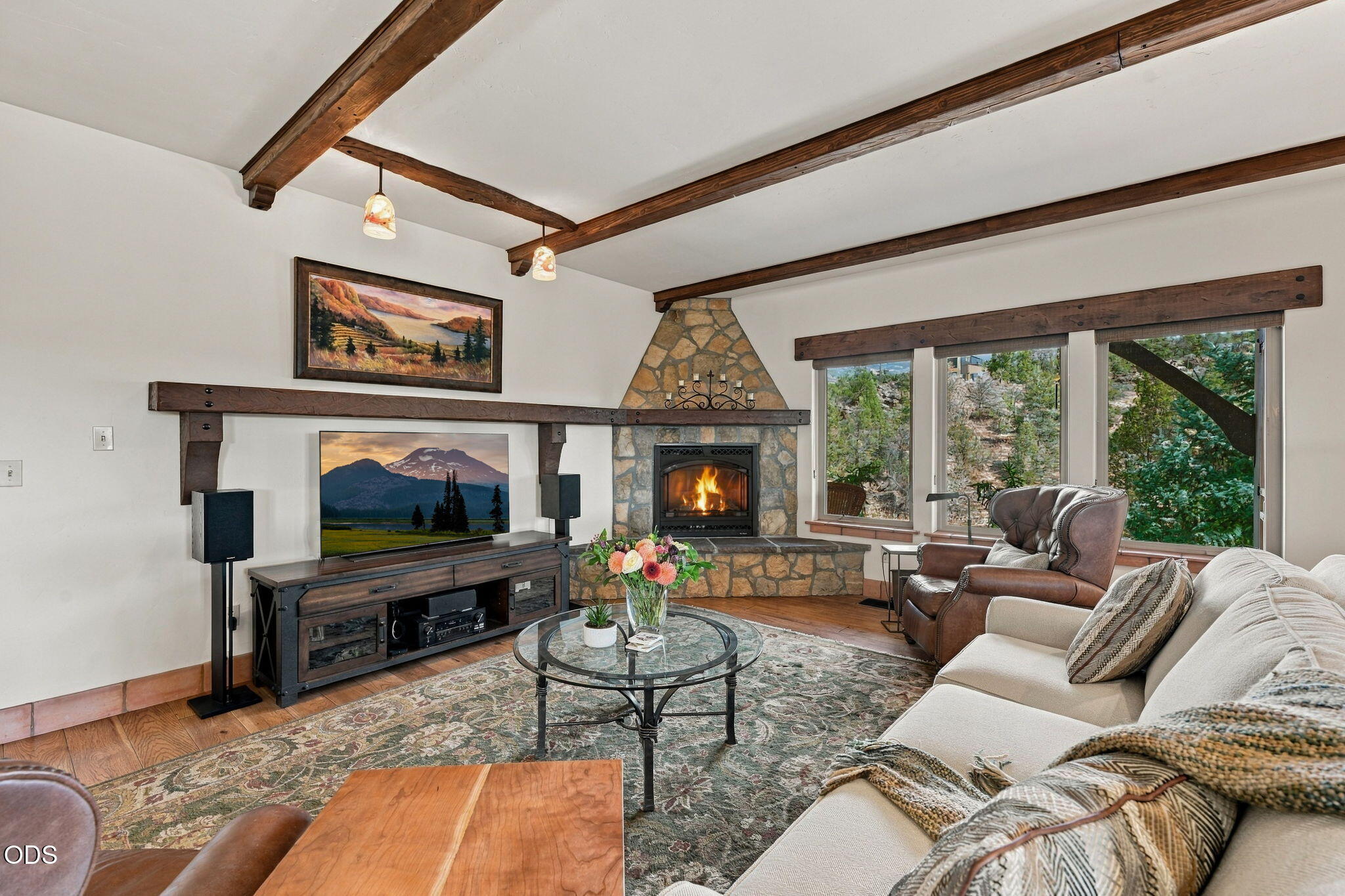 Living room with exposed wood beams, a stone fireplace, and large windows overlooking trees.