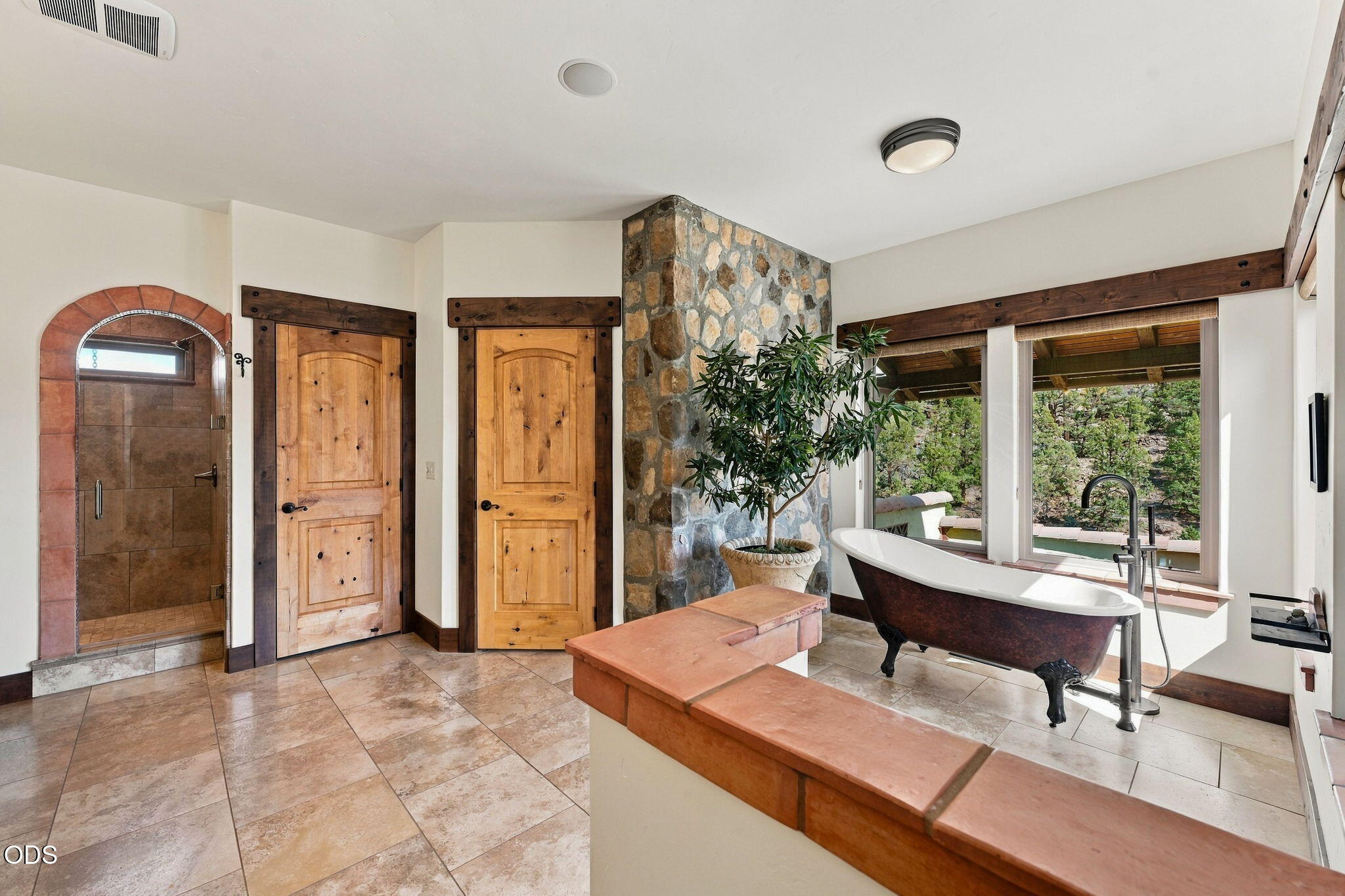 Bathroom with a stone accent wall, tiled shower, and freestanding tub beside windows overlooking trees.