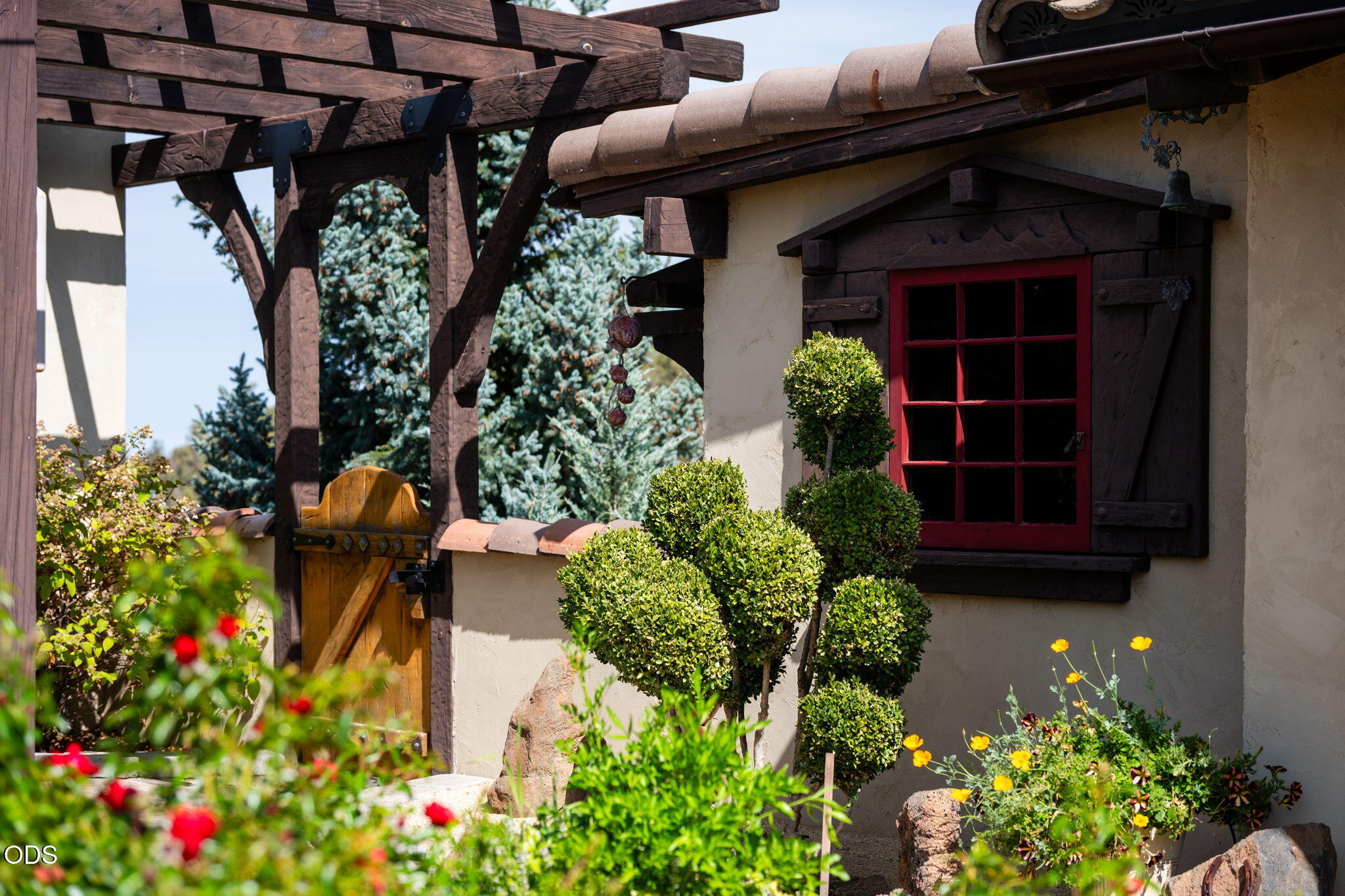 Home exterior with a red-framed window, timber pergola, and sculpted shrubs among flowering plants.