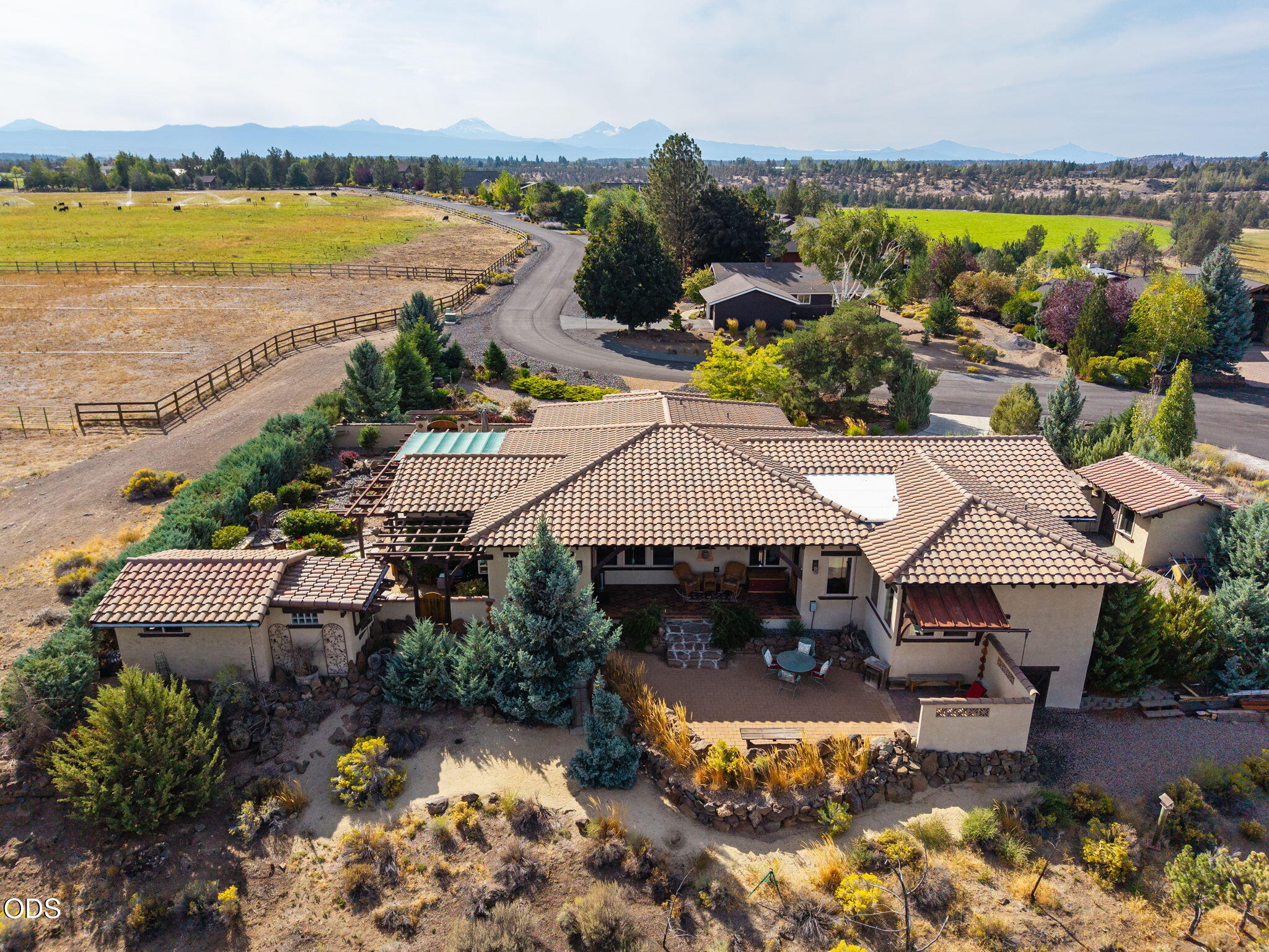 Aerial view of a tile-roofed home with landscaped grounds, fenced pasture, and mountains in the distance.