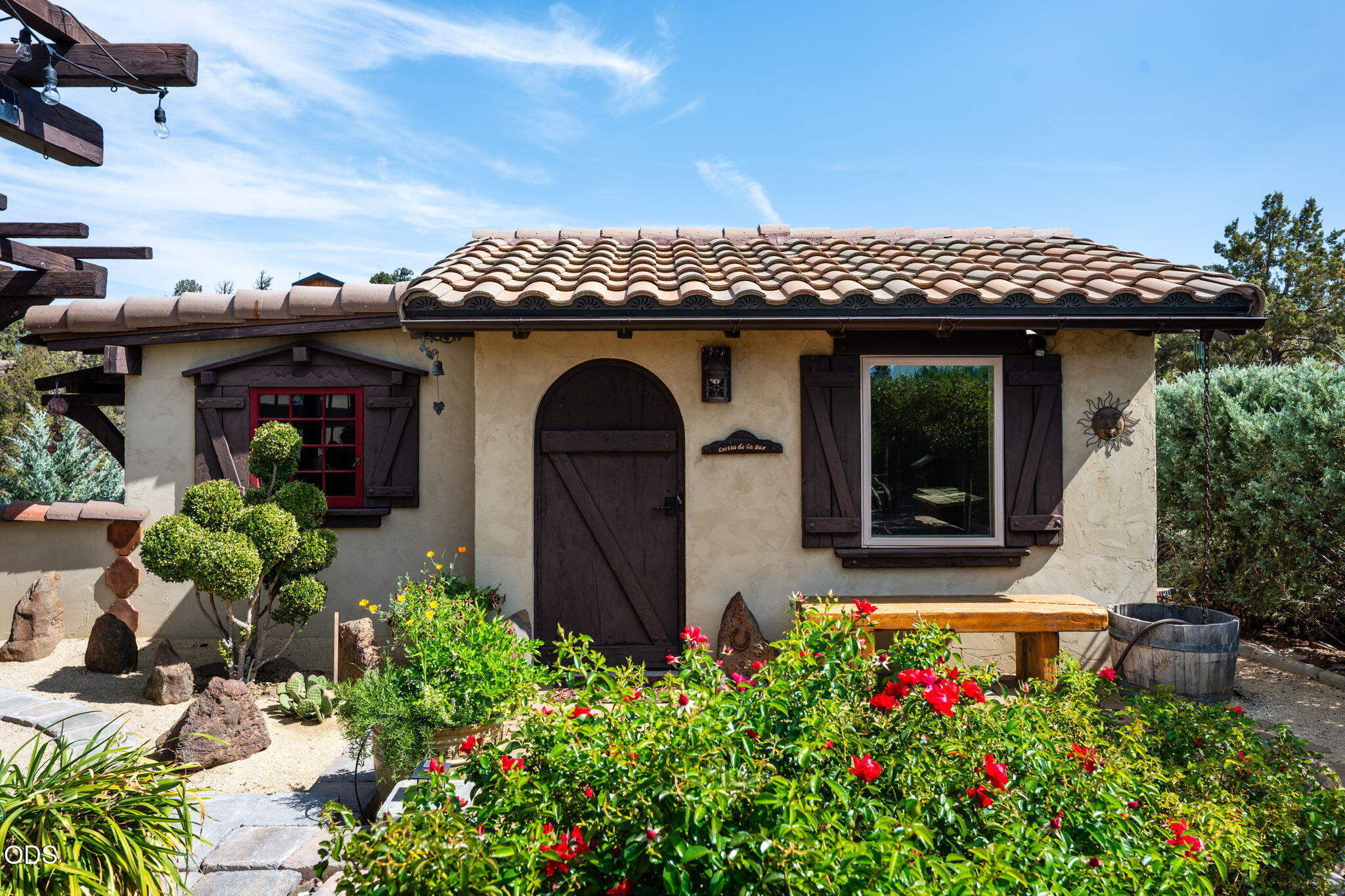 Stucco home with a tile roof, arched wooden door, dark shutters, and flowering front garden.