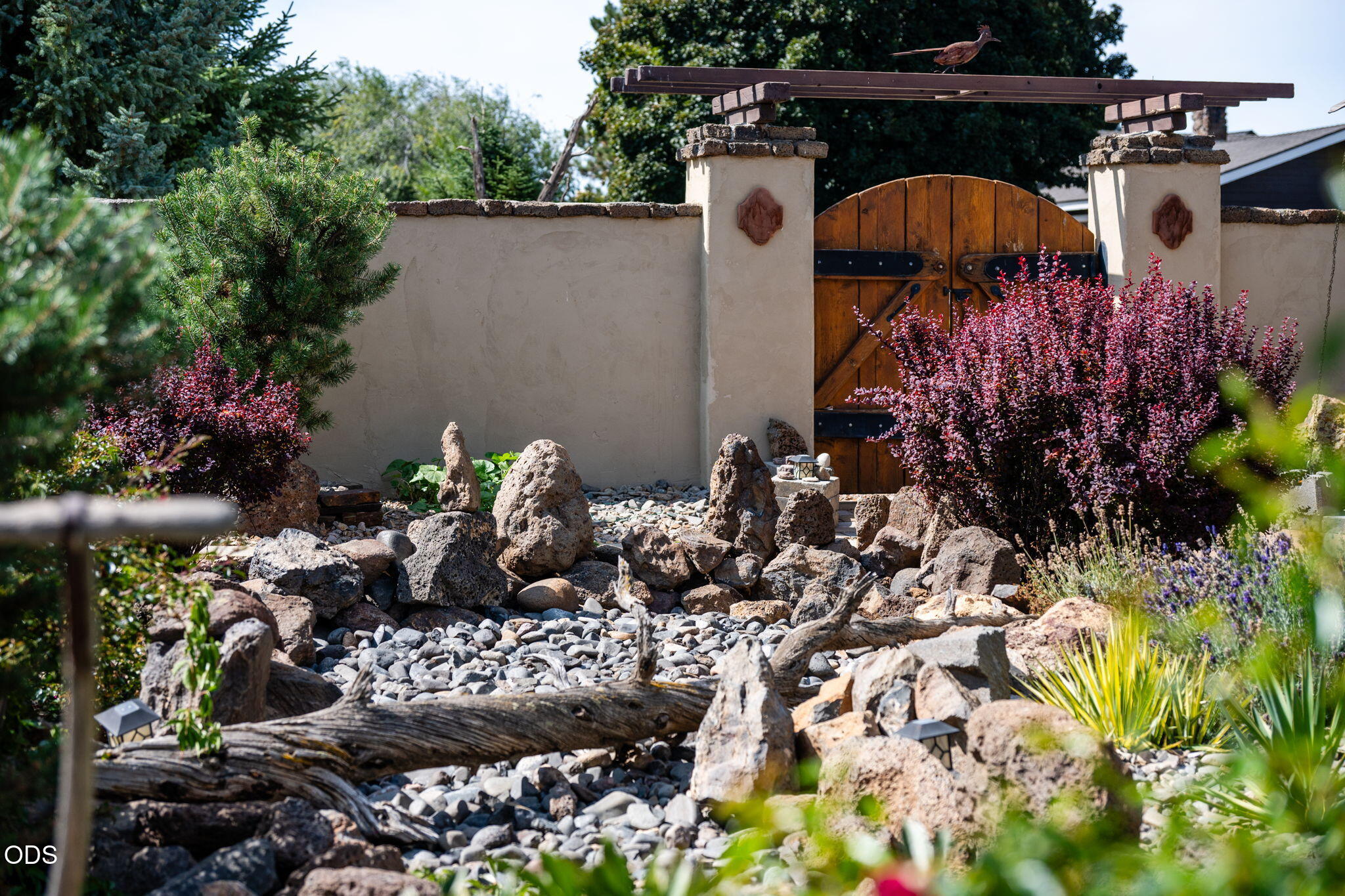 Wooden arched entrance gate above a rock garden with shrubs and flowering plants.