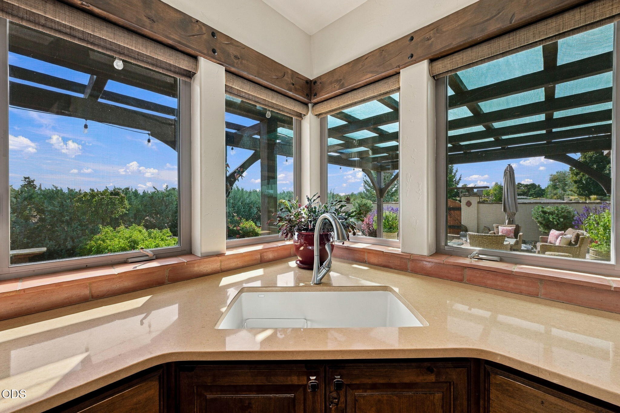 Kitchen sink and pale stone counter beside large windows overlooking a garden and covered patio.