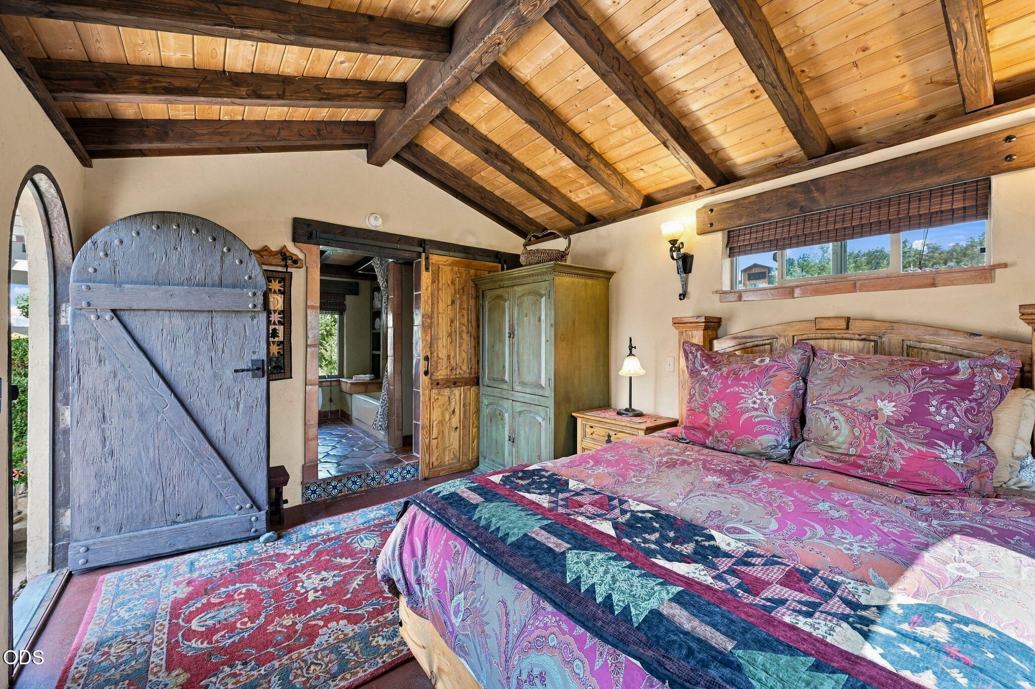 Bedroom with a vaulted timber ceiling, colorful quilt, and large arched barn-style door.