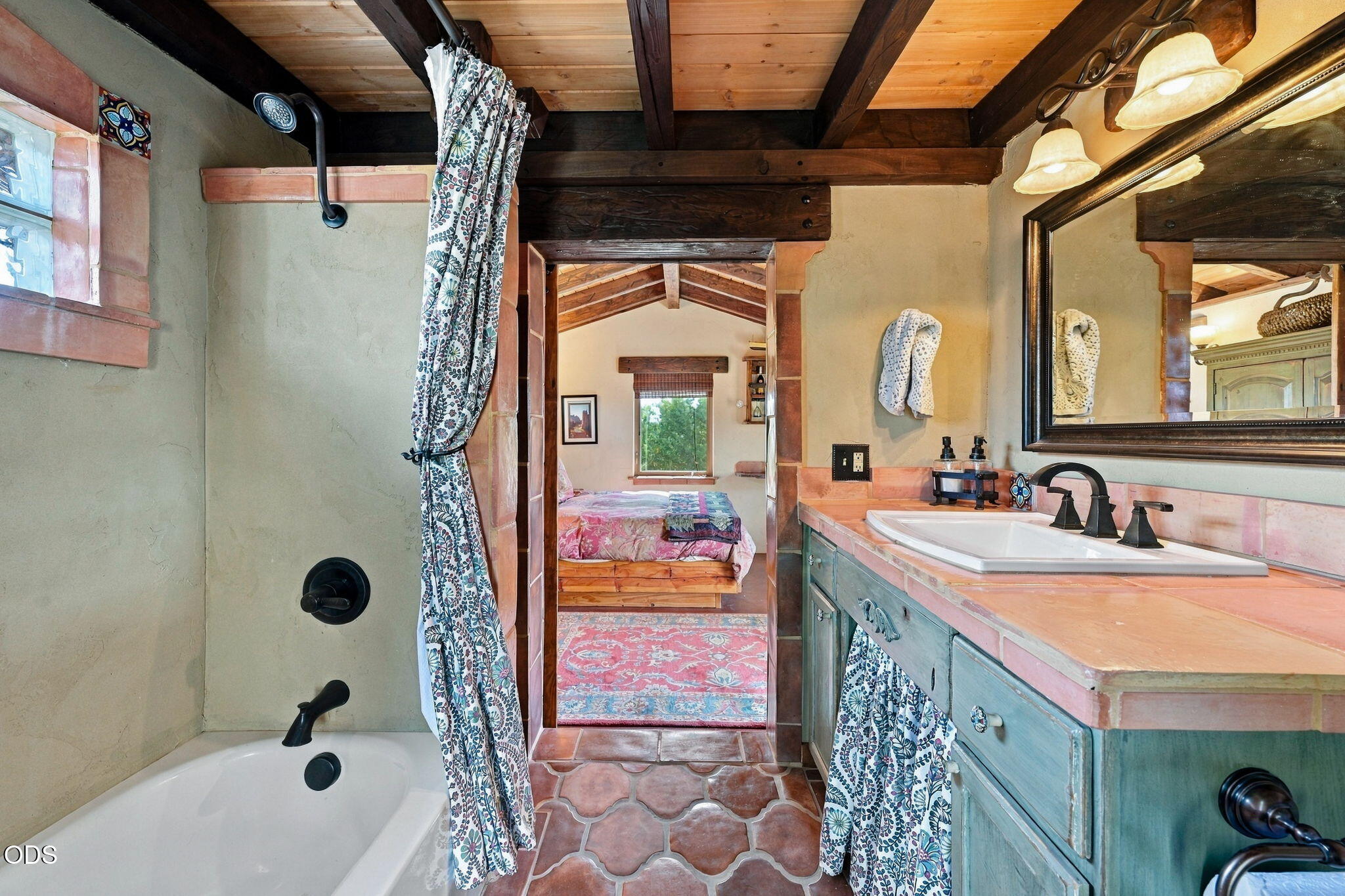Bathroom with a tiled tub and blue-green vanity, looking through to a bedroom.