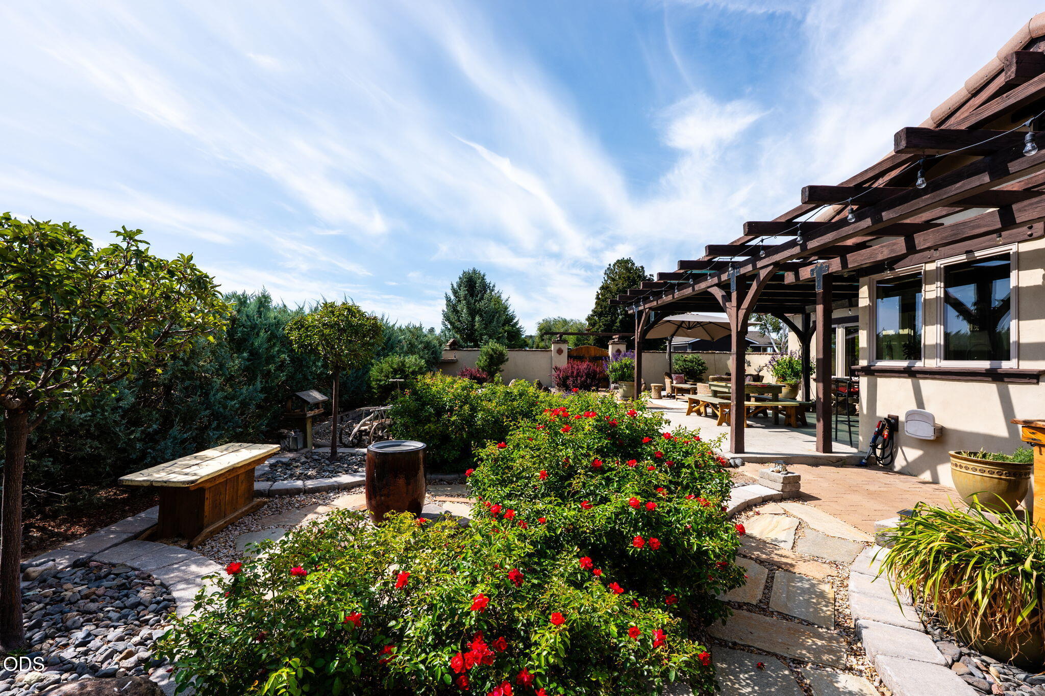 Landscaped backyard patio with a timber pergola, outdoor dining area, stone paths, and red-flowering shrubs.