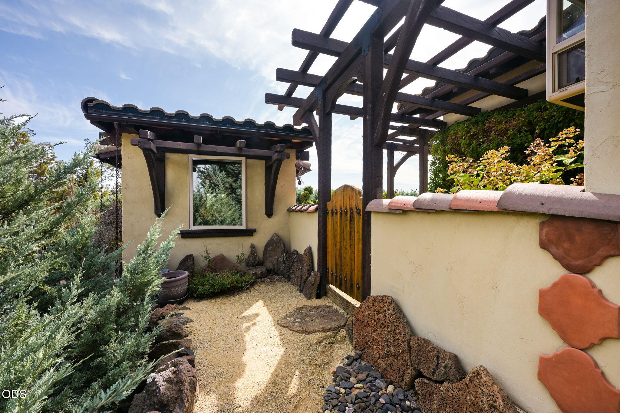 Home entry courtyard with timber pergola, wooden gate, gravel path, and flowering shrubs.