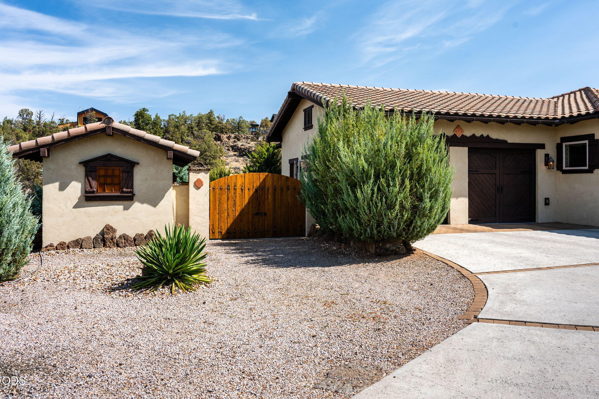 Stucco home entrance with terracotta tile roof, arched wooden gate, gravel landscaping, and curved driveway.
