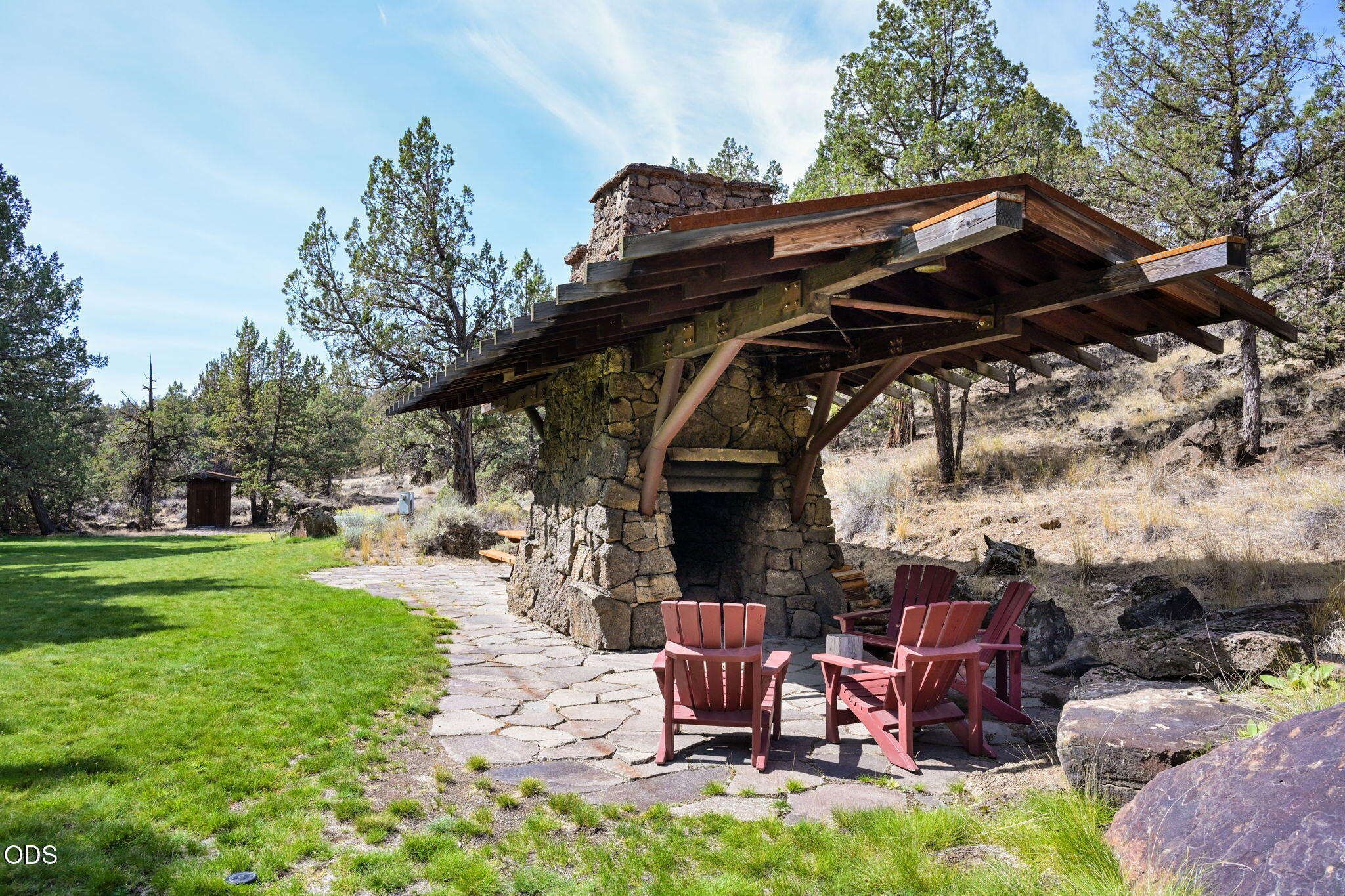 Stone fireplace pavilion with a broad timber roof and red Adirondack chairs on a flagstone patio.