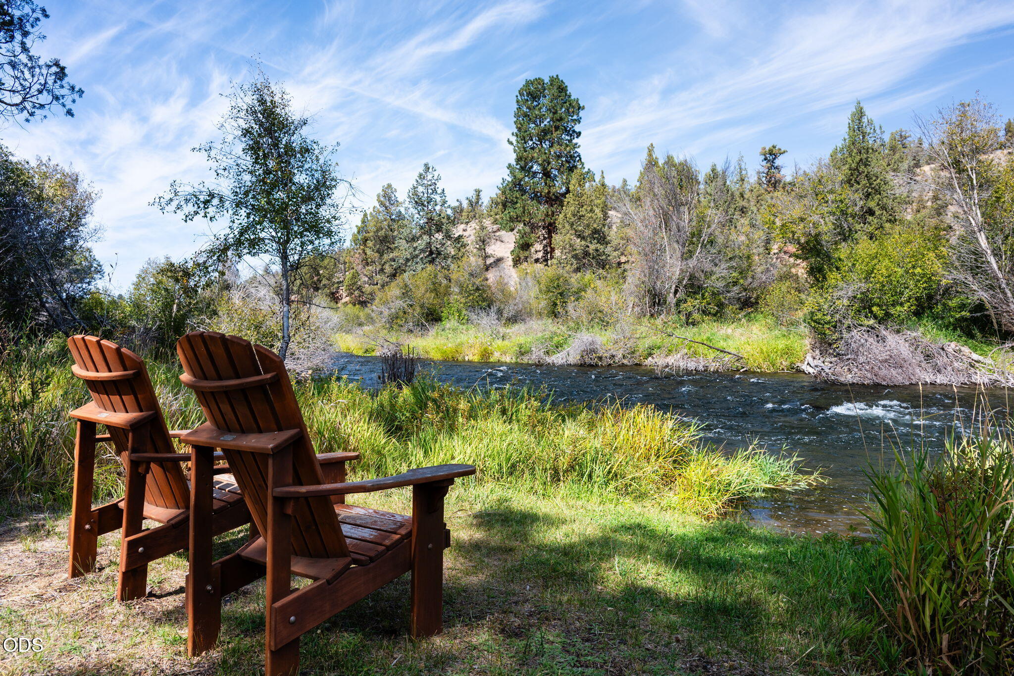 Two wooden Adirondack chairs overlook a flowing river bordered by grasses and trees.