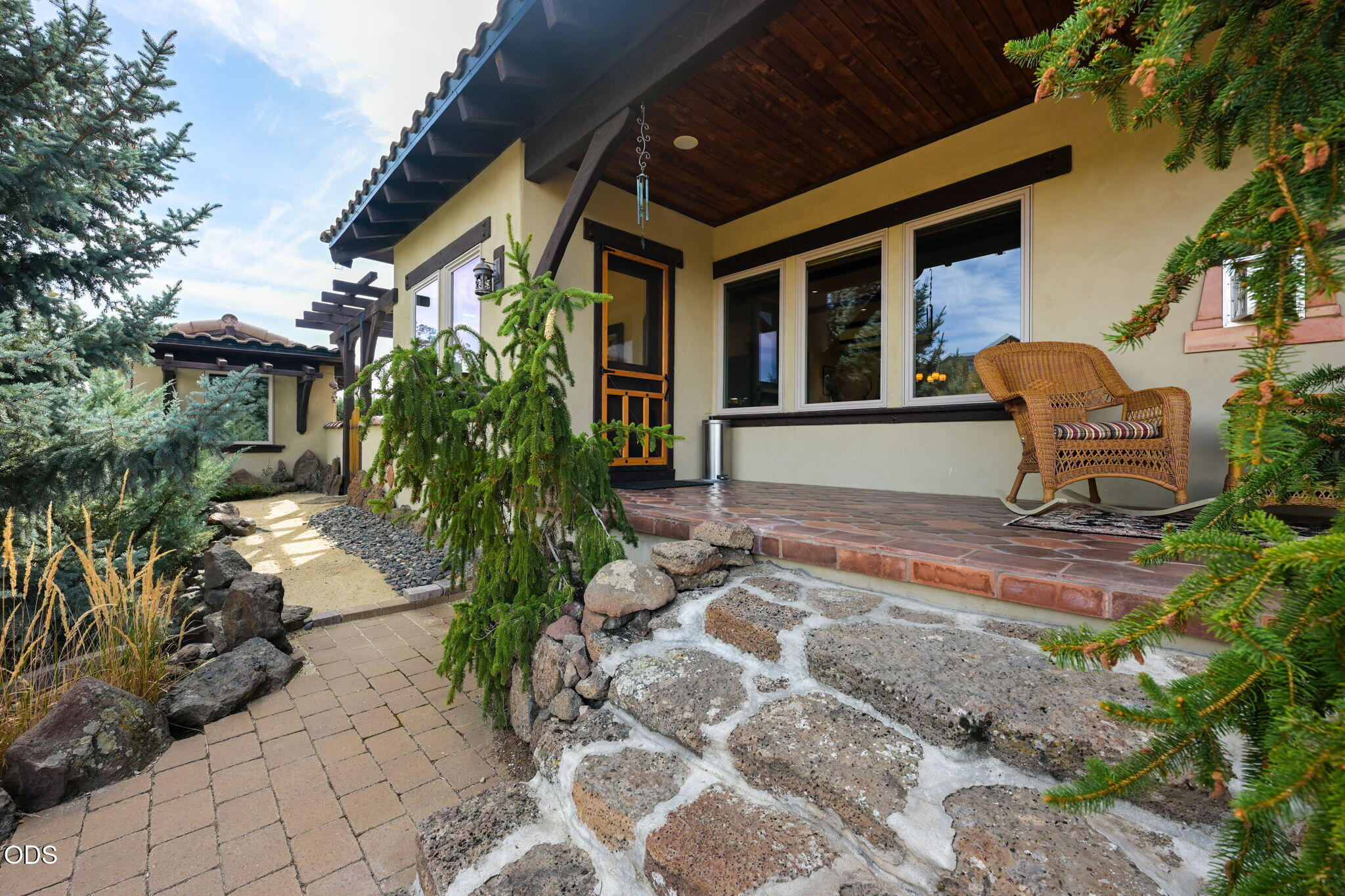 Covered home porch with wicker chair, tiled floor, stone steps, and evergreen landscaping